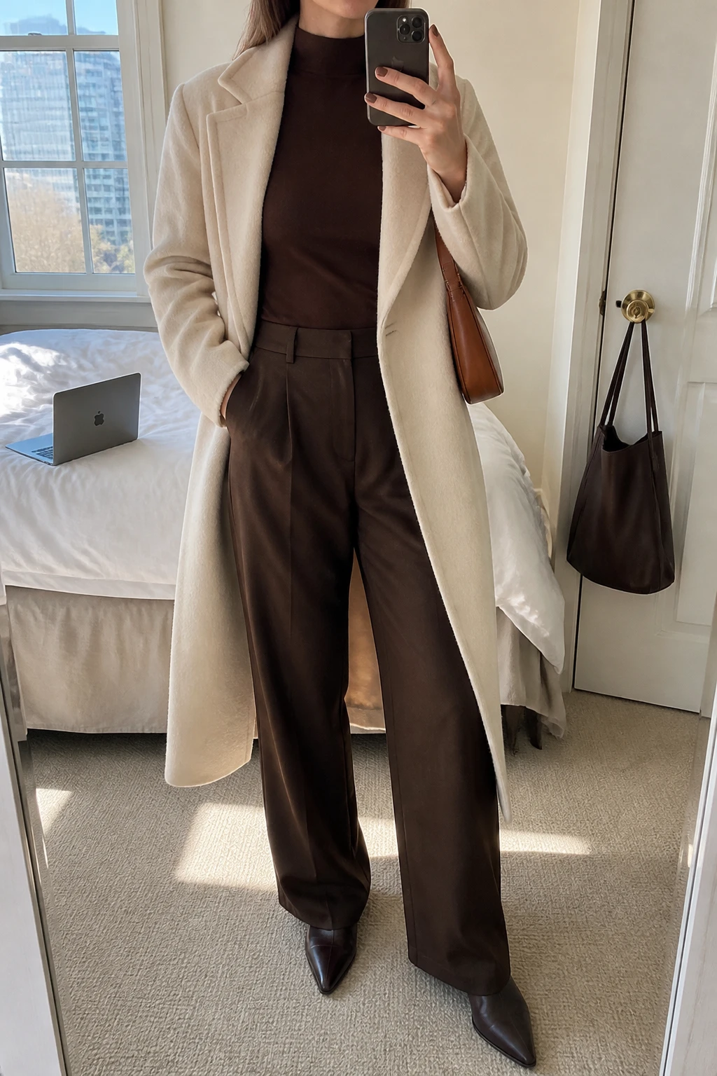 Image Prompt: Mirror selfie of a woman wearing a long cream tailored wool coat open over a chocolate brown fitted fine-knit mock-neck sweater, matching chocolate brown high-waisted wide-leg tailored trousers, dark brown leather pointed-toe ankle boots with low block heels, and a small caramel leather shoulder bag, holding a phone in front of a mirror, cropped at the NECK so the face does not show, with a relaxed casual selfie pose, in a stylish city apartment, soft natural window light, a laptop on the bed and a handbag hanging on a door handle, natural cream wool texture, realistic matching brown fabric folds, latest iPhone photo quality, no additional clothing or accessories.