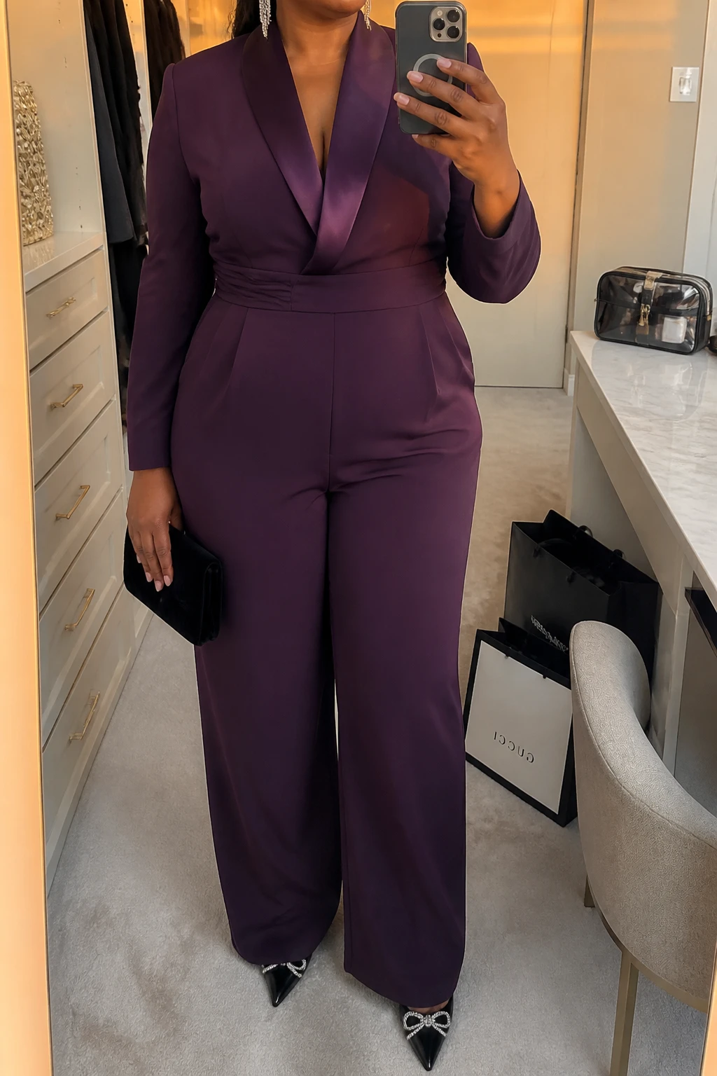 Mirror selfie of a plus size woman wearing a plum purple tuxedo-style wide-leg jumpsuit with satin lapels, long sleeves, and a structured waistband, black pointed pumps with small crystal details, a black velvet clutch, and silver linear earrings, clearly showing the exact jumpsuit, plum color, satin lapels, footwear, and accessories listed, holding a phone in front of a mirror, cropped at the neck so her face does not appear, relaxed confident selfie pose, in a walk-in closet mirror with evening golden-hour lighting, shopping bags beside a chair and a cosmetic bag on the counter, realistic show-stopping graduation outfit, latest iPhone photo quality.