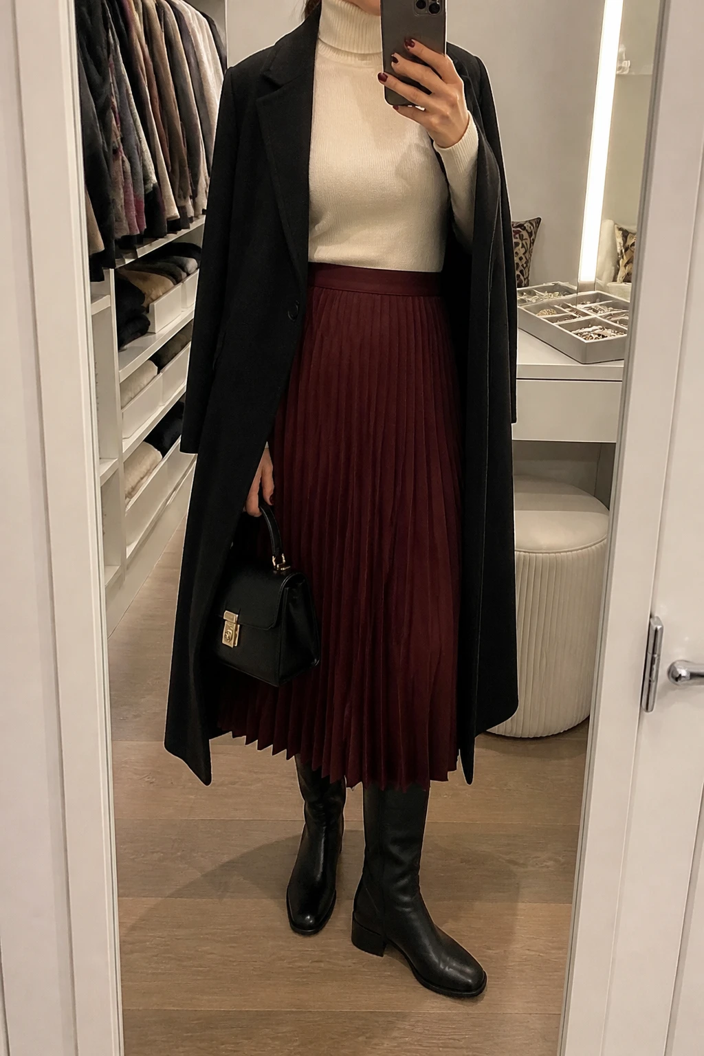 Image Prompt: Mirror selfie of a woman wearing an ivory fitted cashmere-blend turtleneck sweater tucked into a burgundy wool-blend pleated midi skirt, black leather knee-high boots with low block heels, a long black tailored wool coat worn open, and a small black structured top-handle handbag, holding a phone in front of a mirror, cropped at the NECK so the face does not show, with a relaxed casual selfie pose, in a walk-in closet mirror, warm soft indoor lighting, an open closet with clothes visible and a jewelry tray on the vanity, realistic burgundy pleats, natural ivory knit texture and black wool coat drape, latest iPhone photo quality, no additional clothing or accessories.