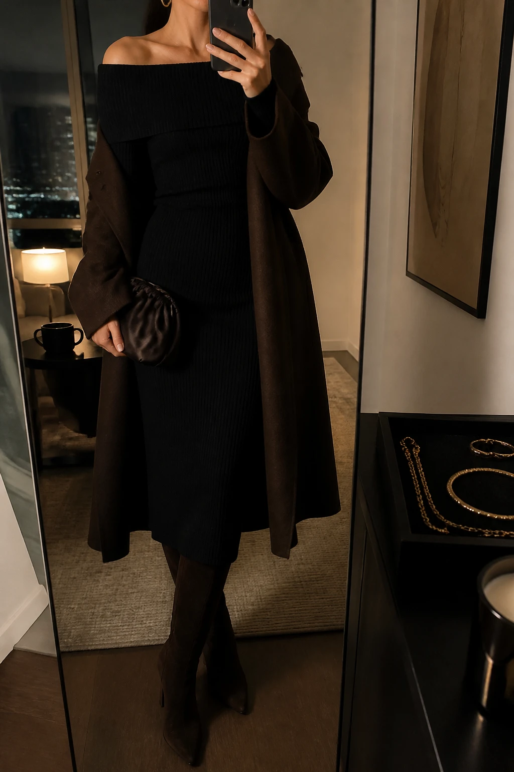 Image Prompt: Mirror selfie of a woman wearing a black long-sleeve off-shoulder ribbed knit midi dress with a folded neckline and softly fitted waist, a long chocolate brown tailored wool coat worn open, dark brown suede knee-high boots with block heels, a small dark brown satin clutch, and delicate gold hoop earrings, holding a phone in front of a mirror, cropped at the NECK so the face does not show, with a relaxed casual selfie pose, in a stylish city apartment, warm moody lighting, a coffee mug on a side table and a jewelry tray on the vanity, realistic black ribbed knit texture, natural chocolate wool coat folds, elegant evening lighting, latest iPhone photo quality, no additional clothing or accessories.