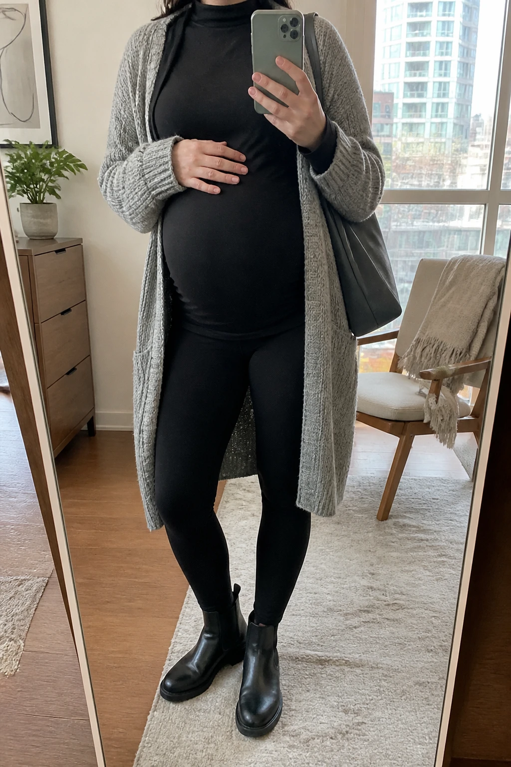 Image Prompt: Mirror selfie of a visibly pregnant woman wearing a long heather grey chunky-knit open-front cardigan over a black long-sleeve maternity tunic with a soft mock neckline and extra fabric draping over her rounded belly, black high-waisted over-the-bump fleece-lined maternity leggings, black leather flat Chelsea boots with rubber soles, and a medium charcoal grey nylon shoulder tote, holding a phone in front of a mirror, cropped at the NECK so the face does not show, with a relaxed casual selfie pose and natural maternity silhouette, in a stylish city apartment, soft natural window light, a small plant on a dresser and a folded blanket on a chair, realistic grey knit texture, natural black leggings fabric, comfortable casual pregnancy styling, latest iPhone photo quality, no additional clothing or accessories.