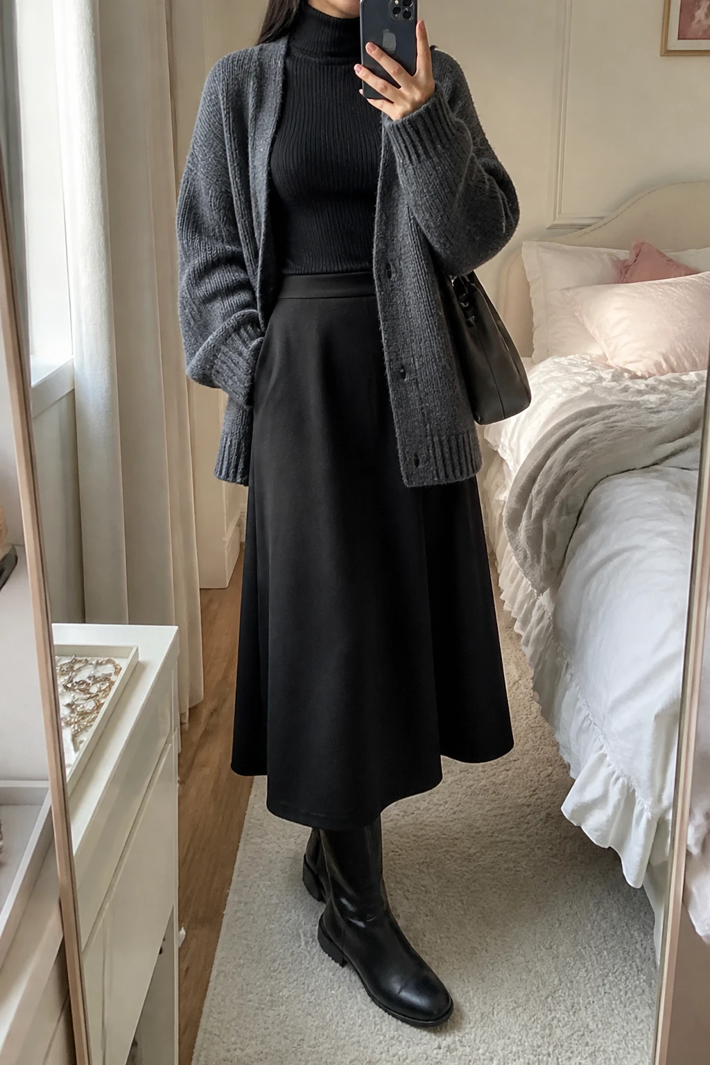 Image Prompt: Mirror selfie of a woman wearing a charcoal grey oversized chunky-knit cardigan over a black fitted ribbed long-sleeve turtleneck top, a black wool-blend A-line maxi skirt, black opaque fleece-lined tights, black leather flat knee-high boots, and a small charcoal grey leather shoulder bag, holding a phone in front of a mirror, cropped at the NECK so the face does not show, with a relaxed casual selfie pose, in a feminine-themed bedroom, natural window light, soft pillows on an unmade bed and a jewelry tray on the vanity, realistic thick cardigan texture, natural skirt folds, latest iPhone photo quality, no additional clothing or accessories.