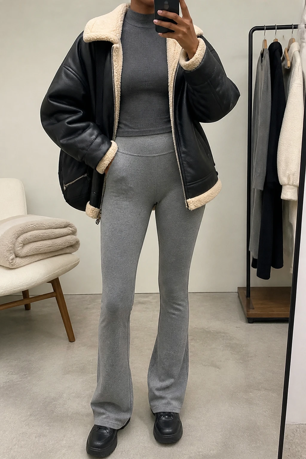 Image Prompt: Mirror selfie of a black woman wearing a black oversized faux-leather shearling-lined jacket with a cream fleece collar over a charcoal grey fitted thermal mock-neck top, heather grey high-waisted fleece-lined flared leggings, black chunky leather sneakers with flat rubber soles, and a medium black nylon zippered backpack, holding a phone in front of a mirror, cropped at the NECK so the face does not show, with a relaxed casual selfie pose, in a minimal dressing room, soft indoor lighting, a folded blanket on a chair and a clothing rack with garments behind, realistic black leather texture, soft cream shearling trim and natural grey legging folds, latest iPhone photo quality, no additional clothing or accessories.