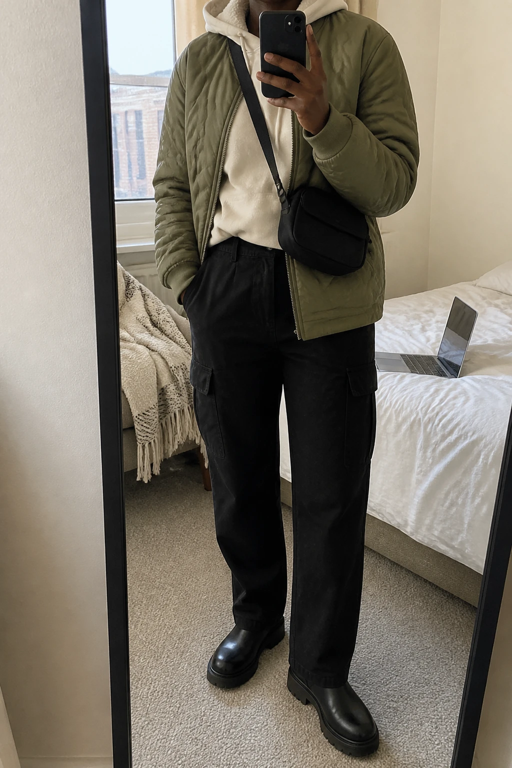 Image Prompt: Mirror selfie of a black woman wearing an olive green insulated quilted hip-length jacket over a cream thick fleece-lined hoodie, black high-waisted relaxed straight-leg cargo trousers, black leather chunky flat Chelsea boots with rubber soles, a small black nylon crossbody bag, and a dark olive ribbed knit beanie, holding a phone in front of a mirror, cropped at the NECK so the face does not show, with a relaxed casual selfie pose, in a cozy bedroom mirror setup, soft natural window light, a laptop on the bed and a folded blanket on a chair, realistic olive quilted fabric, natural cream hoodie folds and practical black cargo trousers, latest iPhone photo quality, no additional clothing or accessories.