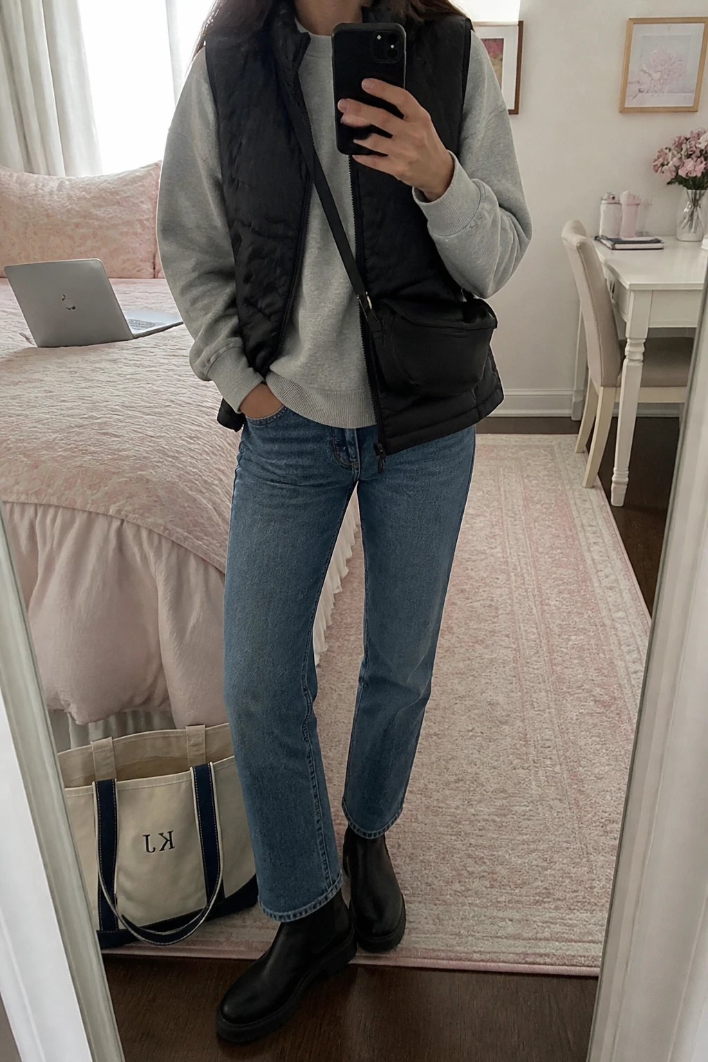Image Prompt: Mirror selfie of a woman wearing a black lightweight quilted insulated puffer vest over a heather grey relaxed fleece-lined crewneck sweatshirt, medium blue straight-leg denim jeans, black leather chunky Chelsea boots with flat rubber soles, and a small black nylon crossbody bag, holding a phone in front of a mirror, cropped at the NECK so the face does not show, with a relaxed casual selfie pose, in a feminine-themed bedroom, soft natural window light, a laptop on the bed and a tote bag resting on the floor, realistic black quilted fabric, natural grey fleece texture and everyday denim folds, latest iPhone photo quality, no additional clothing or accessories.