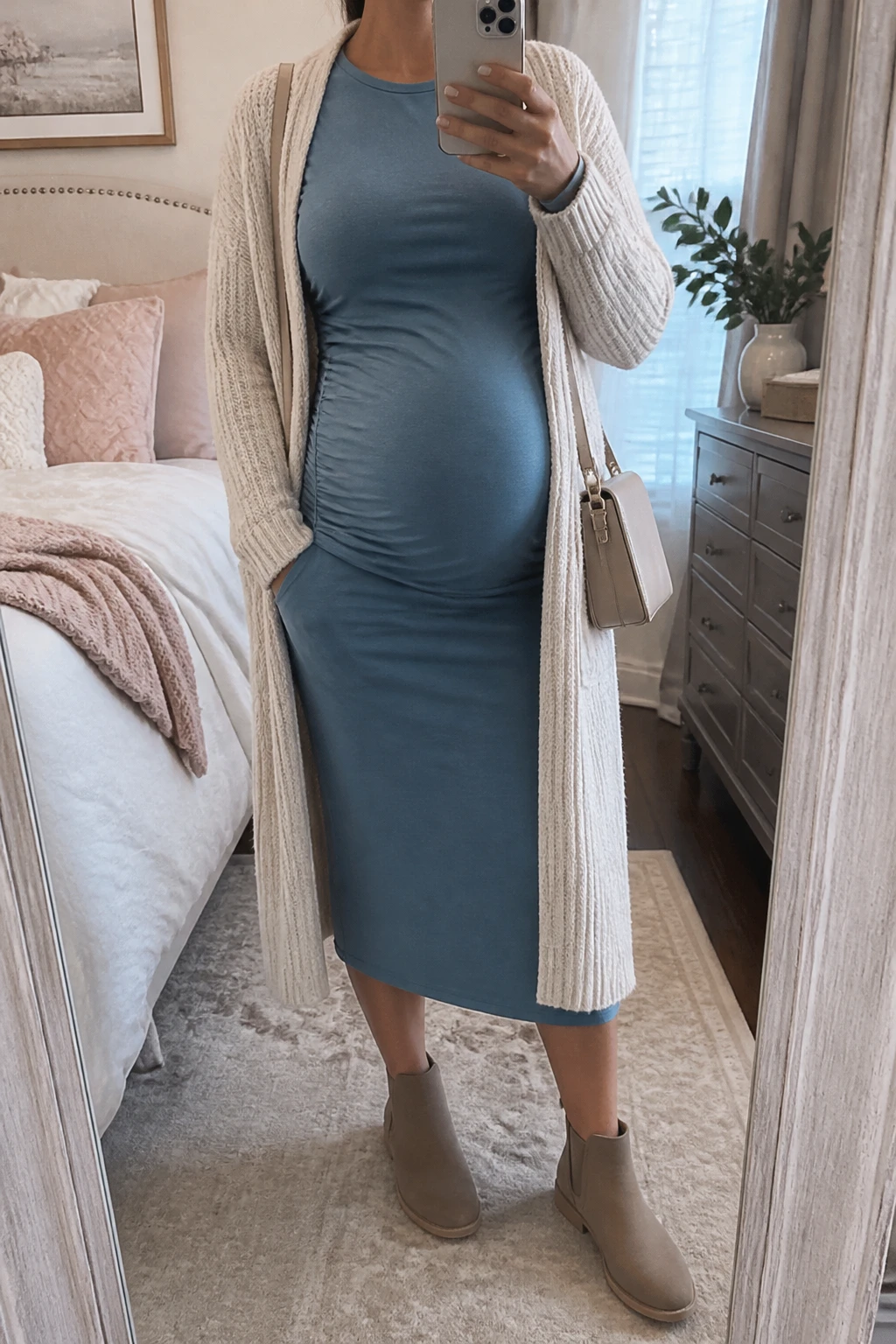 Image Prompt: Mirror selfie of a visibly pregnant woman wearing a dusty blue long-sleeve soft jersey maternity midi dress with a modest round neckline and gentle side gathering around her growing baby bump, a cream longline chunky-knit open-front cardigan, taupe suede flat ankle boots with rubber soles, a small beige leather crossbody bag, and delicate silver stud earrings, holding a phone in front of a mirror, cropped at the NECK so the face does not show, with a relaxed casual selfie pose and realistic pregnant proportions, in a feminine-themed bedroom, soft natural window light, soft pillows on an unmade bed and a small plant on a dresser, realistic dusty blue jersey fabric, natural cream cardigan folds, soft muted winter colors, latest iPhone photo quality, no additional clothing or accessories.