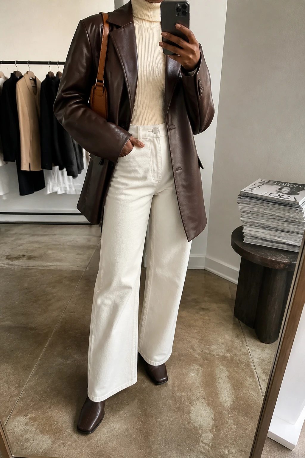 Image Prompt: Mirror selfie of a black woman wearing an oversized chocolate brown faux-leather single-breasted blazer over a cream fitted ribbed turtleneck sweater, winter-white high-waisted wide-leg denim jeans, dark brown leather square-toe ankle boots with low block heels, and a small caramel leather shoulder bag, holding a phone in front of a mirror, cropped at the NECK so the face does not show, with a relaxed casual selfie pose, in a fashion studio mirror corner, soft indoor lighting, a clothing rack with garments behind and a stack of fashion magazines on a side table, realistic chocolate leather grain, natural cream denim folds, warm natural brown skin visible on hands, latest iPhone photo quality, no additional clothing or accessories.