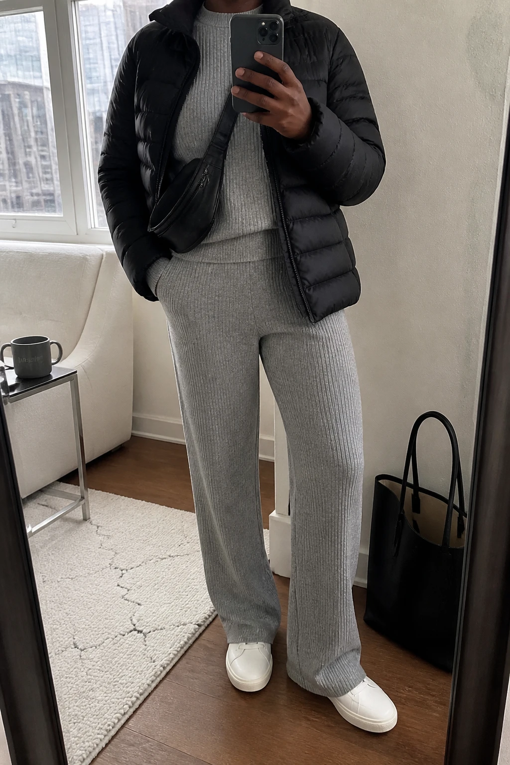 Image Prompt: Mirror selfie of a black woman wearing a matching heather grey soft ribbed knit lounge set consisting of a relaxed crewneck sweater and high-waisted straight-leg trousers, a black lightweight insulated quilted puffer jacket worn open, white leather slip-on sneakers with thick cream rubber soles, and a medium black nylon zippered crossbody bag, holding a phone in front of a full-length mirror, cropped at the NECK so the face does not show, with a relaxed casual selfie pose, in a stylish city apartment, soft natural window light, a tote bag resting on the floor and a coffee mug on a side table, realistic soft knit texture, natural trouser folds, slightly imperfect everyday travel styling, latest iPhone photo quality, no additional clothing or accessories.