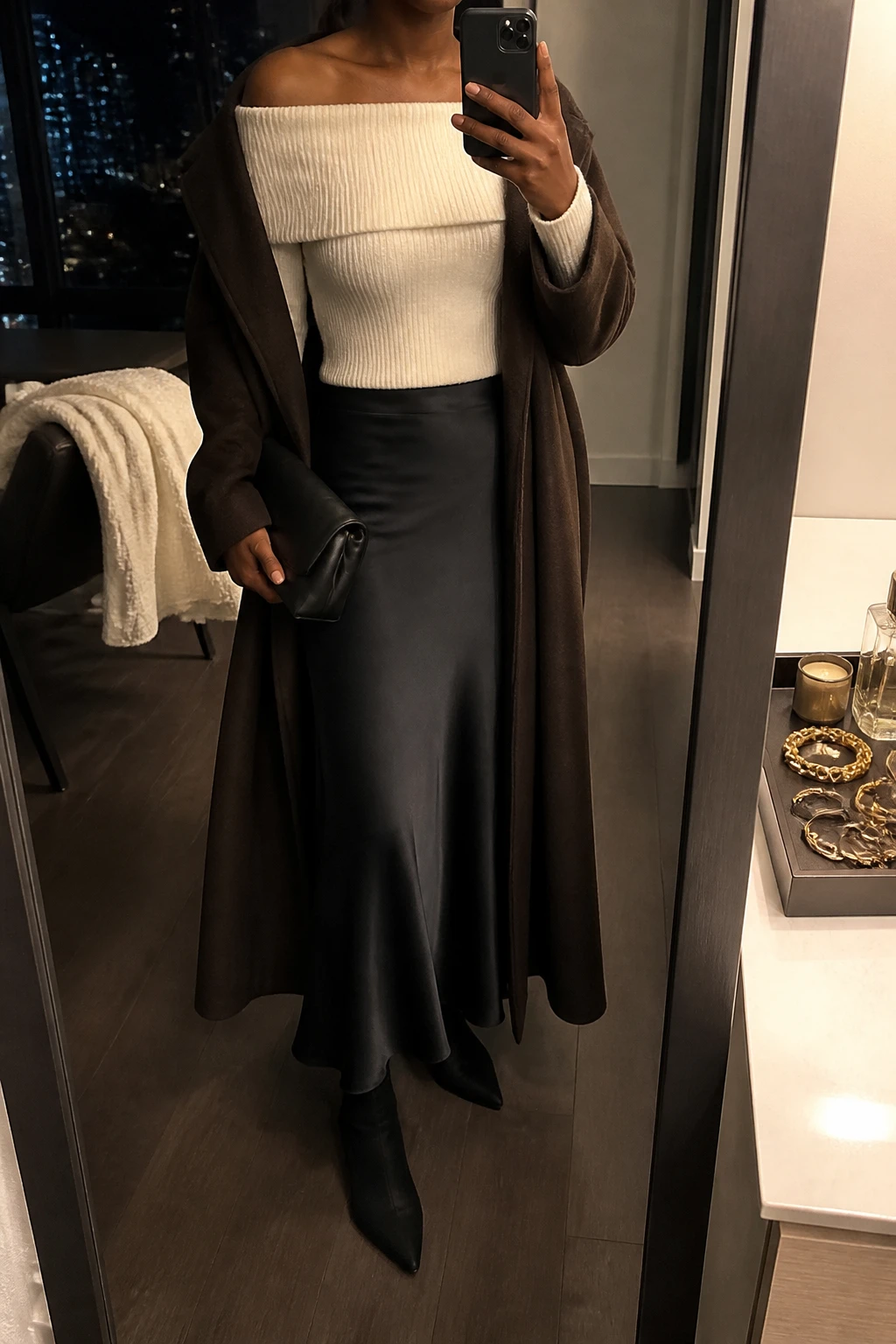 Image Prompt: Mirror selfie of a black woman wearing an ivory fitted off-shoulder ribbed knit sweater with a wide folded neckline tucked into a black satin bias-cut maxi skirt, black suede pointed-toe ankle boots with low block heels, a long chocolate brown tailored wool coat worn open, and a small black satin clutch, holding a phone in front of a mirror, cropped at the NECK so the face does not show, with a relaxed casual selfie pose, in a stylish city apartment, warm moody lighting, a jewelry tray on the vanity and a folded blanket on a chair, realistic ivory ribbed knit texture, natural black satin reflections and elegant evening fabric drape, latest iPhone photo quality, no additional clothing or accessories.