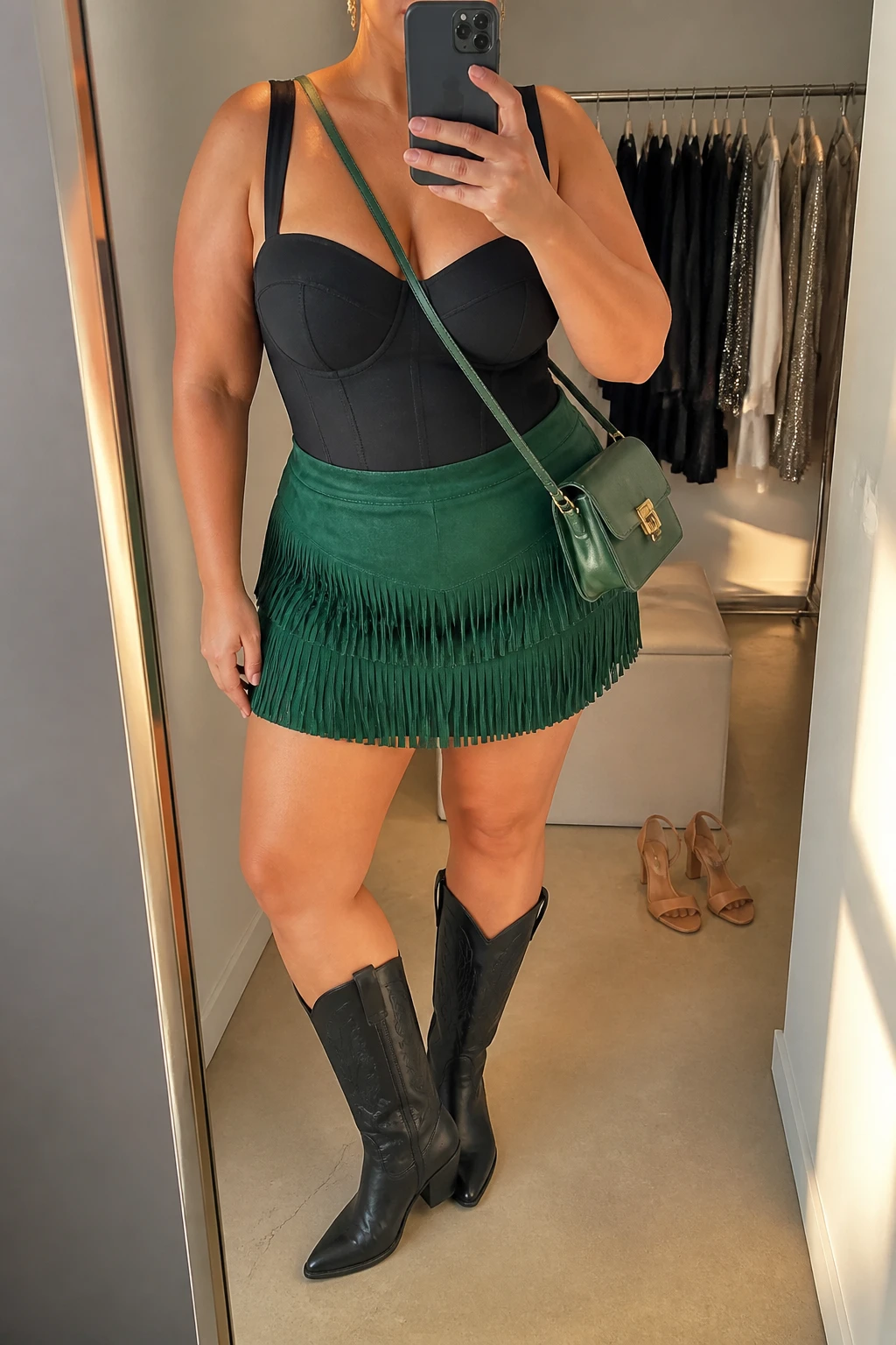 Mirror selfie of a plus size woman wearing an emerald green suede mini skirt with layered fringe, a structured black corset-style bodysuit with wide supportive straps, black knee-high cowboy boots with low block heels, a small emerald green leather crossbody bag, and gold drop earrings, clearly showing the exact clothing items, emerald suede fringe, black structured top, colors, footwear, and accessories listed, holding a phone in front of a mirror, cropped at the neck so the face is hidden, relaxed energetic selfie pose, in a minimal dressing room with evening golden-hour lighting, a clothing rack with garments behind her and a pair of heels on the floor nearby, realistic bold night-out outfit, latest iPhone photo quality.