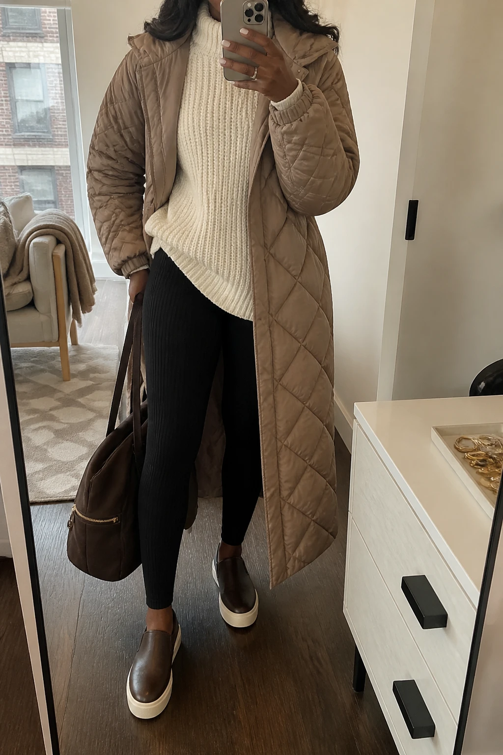 Image Prompt: Mirror selfie of a black woman wearing a cream oversized chunky-knit mock-neck sweater that extends below the hips, black high-waisted thick ribbed thermal leggings, a long taupe lightweight quilted puffer coat worn open, chocolate brown cushioned leather slip-on sneakers with cream soles, and a large dark brown nylon zippered tote bag with sturdy shoulder straps, holding a phone in front of a mirror, cropped at the NECK so the face does not show, with a relaxed casual selfie pose, in a stylish city apartment, soft natural window light, a folded blanket on a chair and a jewelry tray on the vanity, realistic cream knit stitches, natural taupe puffer texture and comfortable everyday travel proportions, latest iPhone photo quality, no additional clothing or accessories.