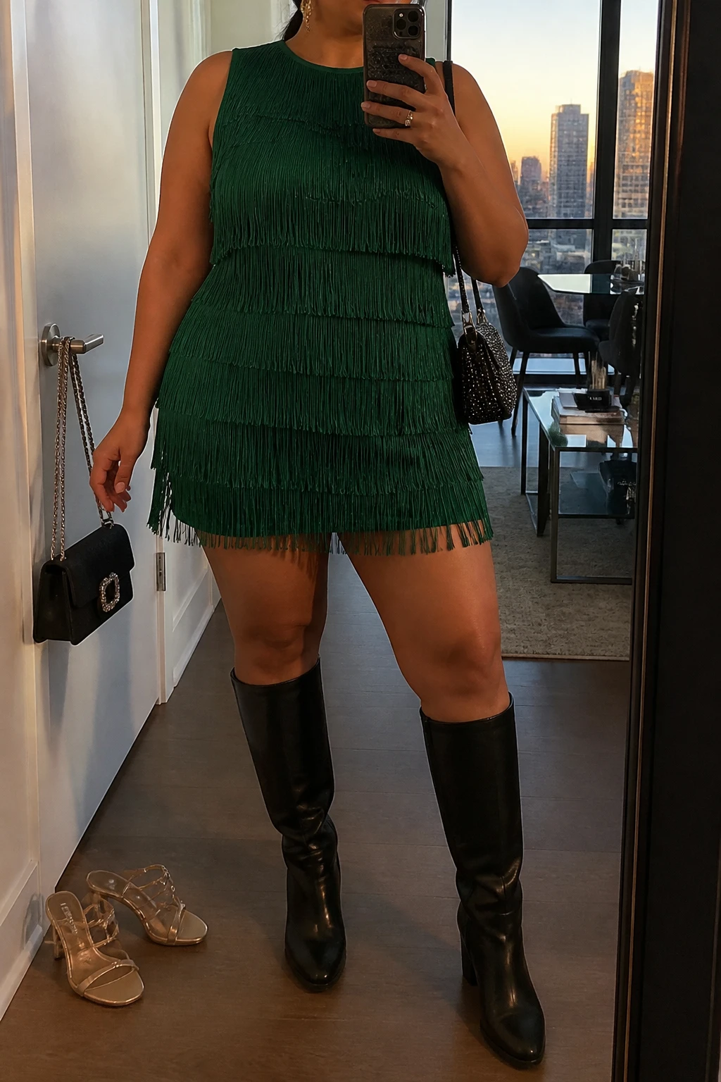 Mirror selfie of a plus size woman wearing an emerald green sleeveless mini dress with layered fringe and a fitted opaque lining, black leather knee-high boots with low block heels, a small black crystal shoulder bag, and gold drop earrings, clearly showing the exact dress, emerald fringe, black lining, footwear, and accessories listed, holding a phone in front of a mirror, cropped at the neck so the face does not show, relaxed energetic selfie stance, in a stylish city apartment with evening golden-hour lighting, a handbag hanging on a door handle and a pair of heels on the floor nearby, realistic dance-ready birthday outfit, latest iPhone photo quality.