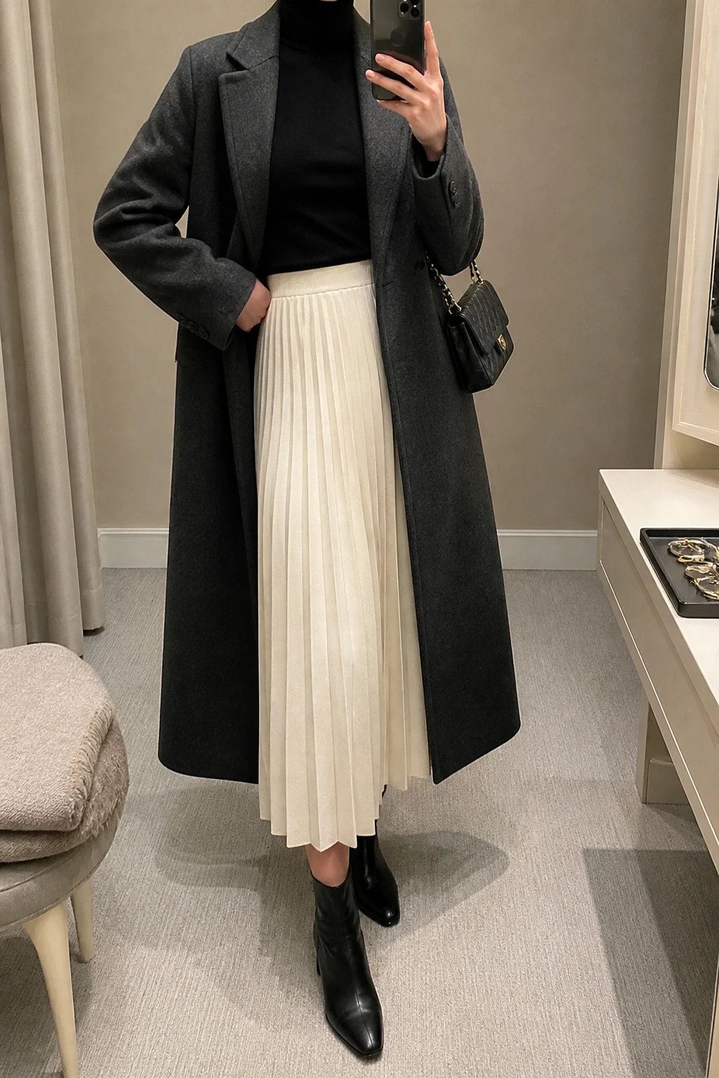 Image Prompt: Mirror selfie of a woman wearing a fitted black cashmere-blend turtleneck tucked into a cream pleated wool-blend midi skirt, black leather heeled ankle boots, a long charcoal grey tailored wool coat worn open, and a small black quilted shoulder bag, holding a phone in front of a mirror, cropped at the NECK so the face does not show, with a relaxed casual selfie pose, in a minimal dressing room, soft indoor lighting, a jewelry tray on the vanity and a folded blanket on a chair, realistic pleated skirt movement and fabric texture, latest iPhone photo quality, no additional clothing or accessories.