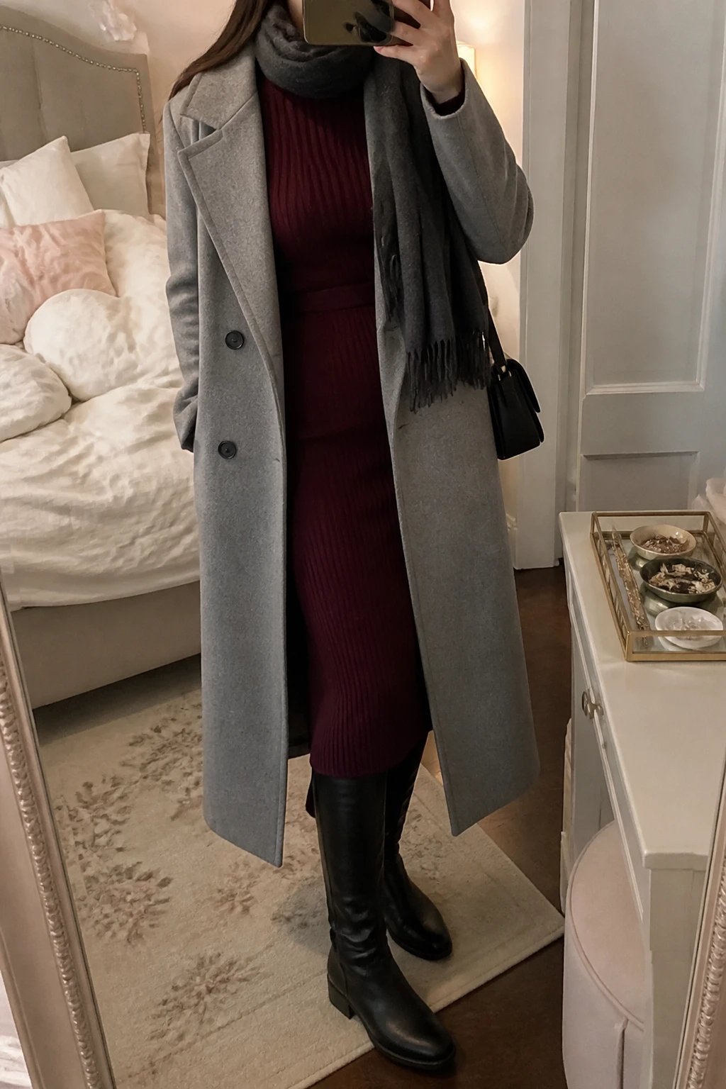Image Prompt: Mirror selfie of a woman wearing a long heather grey tailored wool coat open over a burgundy long-sleeve ribbed knit midi dress with a fitted waist, black leather knee-high low block-heel boots, a small black structured leather shoulder bag, and a charcoal grey cashmere scarf, holding a phone in front of a mirror, cropped at the NECK so the face does not show, with a relaxed casual selfie pose, in a feminine-themed bedroom, warm soft indoor lighting, soft pillows on an unmade bed and a jewelry tray on the vanity, realistic burgundy knit fabric, natural grey coat folds and understated elegant styling, latest iPhone photo quality, no additional clothing or accessories.
