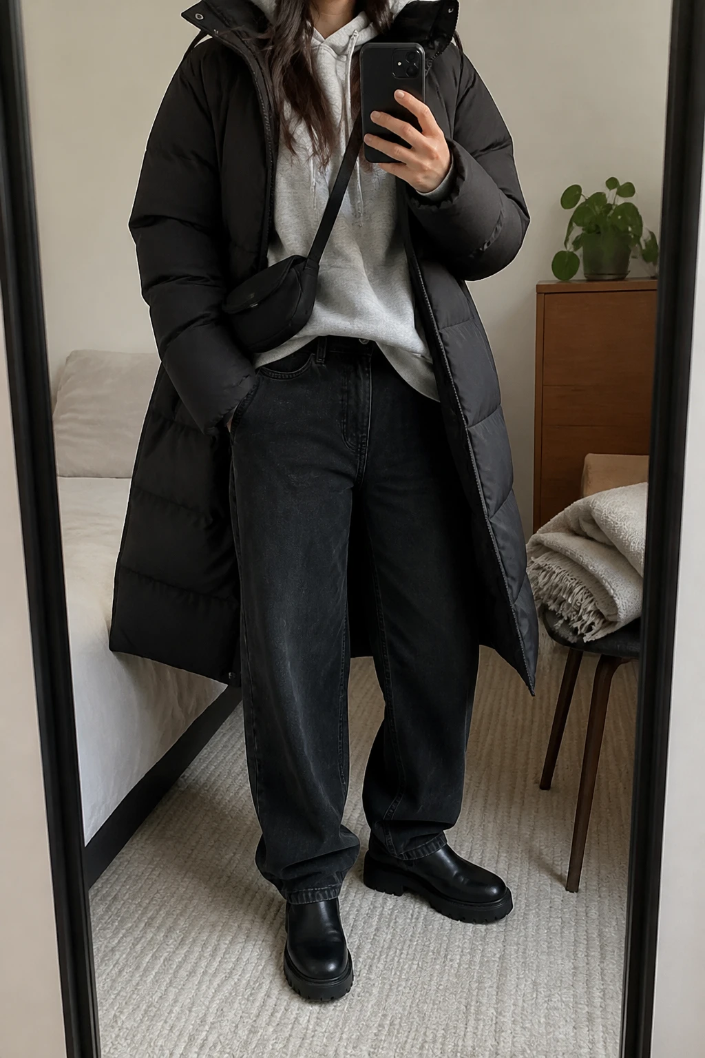 Image Prompt: Mirror selfie of a woman wearing a long black insulated quilted puffer coat over a heather grey oversized fleece-lined hoodie, washed black high-waisted baggy straight-leg denim jeans, black waterproof leather chunky Chelsea boots with thick rubber soles, and a small black nylon crossbody bag, holding a phone in front of a mirror, cropped at the NECK so the face does not show, with a relaxed casual selfie pose, in a cozy bedroom mirror setup, soft indoor lighting, a folded blanket on a chair and a small plant on a dresser, realistic black puffer volume, natural grey hoodie folds and casual black denim texture, latest iPhone photo quality, no additional clothing or accessories.