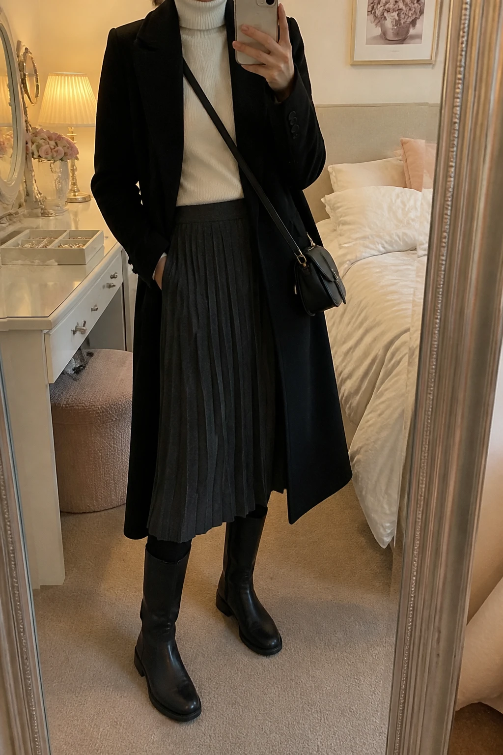Image Prompt: Mirror selfie of a woman wearing a long black tailored wool coat over a cream fitted cashmere turtleneck sweater, a charcoal grey wool-blend pleated midi skirt, black opaque fleece-lined tights, black waterproof leather knee-high boots with low block heels and rubber soles, and a small structured black leather zippered crossbody bag, holding a phone in front of a mirror, cropped at the NECK so the face does not show, with a relaxed casual selfie pose, in a feminine-themed bedroom, warm soft indoor lighting, a jewelry tray on the vanity and soft pillows on an unmade bed, realistic pleated wool fabric, natural cashmere knit and practical winter footwear, latest iPhone photo quality, no additional clothing or accessories.