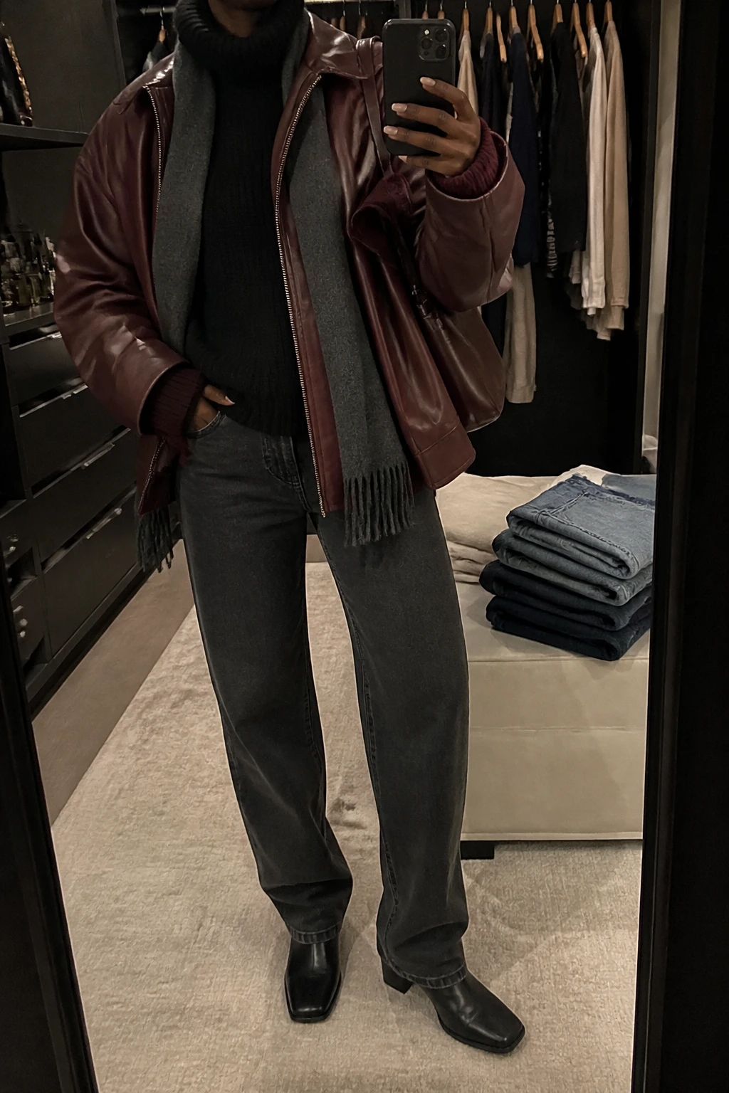 Image Prompt: Mirror selfie of a black woman wearing a burgundy oversized faux-leather jacket over a black heavyweight ribbed turtleneck sweater, washed black straight-leg jeans, black leather square-toe ankle boots with low block heels, a small burgundy leather shoulder bag, and a charcoal grey wool scarf, holding a phone in front of a mirror, cropped at the NECK so the face does not show, with a relaxed casual selfie pose, in a walk-in closet mirror, warm moody indoor lighting, an open closet with clothes visible and folded denim jeans on a bench, natural leather texture, slightly imperfect casual pose, latest iPhone photo quality, no additional clothing or accessories.