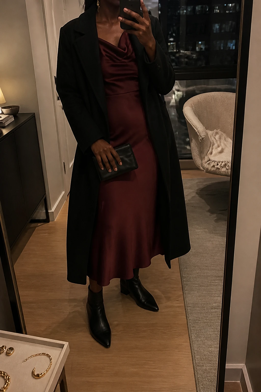 Image Prompt: Mirror selfie of a black woman wearing a deep burgundy long-sleeve satin midi dress with a softly draped cowl neckline and gently fitted waist, black leather pointed-toe ankle boots with low block heels, a long black tailored wool coat worn open, a small black satin evening clutch, and delicate gold drop earrings, holding a phone in front of a full-length mirror, cropped at the NECK so the face does not show, with a relaxed casual selfie pose, in a stylish city apartment, warm moody lighting, a jewelry tray on the vanity and a folded blanket on a chair, realistic burgundy satin draping, natural black wool folds, subtly muted evening colors, natural imperfections, latest iPhone photo quality, no additional clothing or accessories.