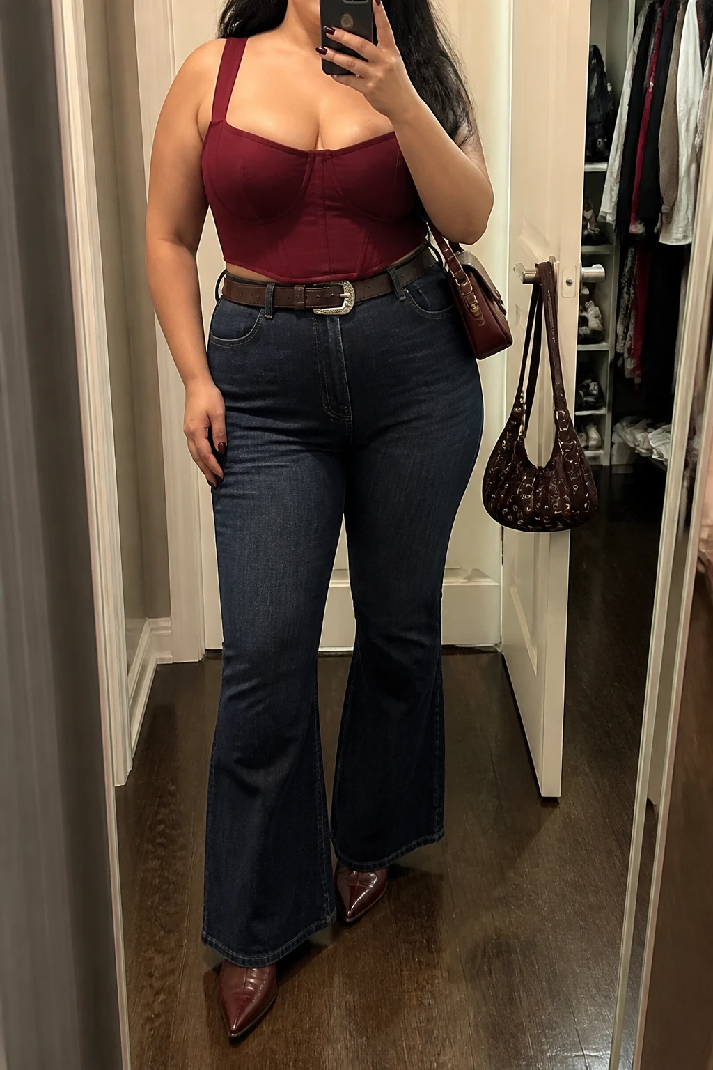 Mirror selfie of a plus size woman wearing dark indigo high-waisted flared jeans, a structured burgundy corset-style top with wide supportive straps, pointed burgundy leather cowboy boots, a dark brown Western belt with a silver buckle, and a small burgundy crossbody bag, clearly showing the exact clothing items, dark denim, burgundy top, matching boots, belt, and accessories listed, holding a phone in front of a mirror, cropped at the neck so her face does not appear, relaxed powerful selfie stance, in a walk-in closet mirror with soft indoor lighting, an open closet with clothes visible and a handbag hanging on a door handle, realistic night-out styling, latest iPhone photo quality.