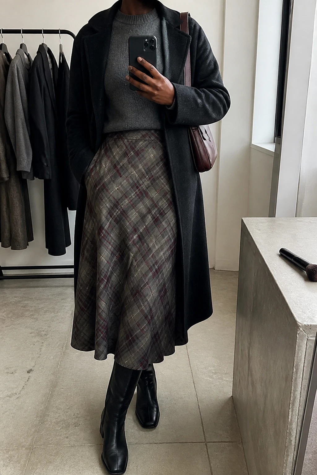 Image Prompt: Mirror selfie of a black woman wearing a charcoal grey fine-knit crewneck sweater tucked into a grey and burgundy plaid wool midi skirt, black leather knee-high low block-heel boots, a black fitted wool coat worn open, and a small burgundy leather shoulder bag, holding a phone in front of a mirror, cropped at the NECK so the face does not show, with a relaxed casual selfie pose, in a minimal dressing room, soft natural window light, a clothing rack with garments behind and a makeup brush on the counter, realistic plaid fabric and natural skirt folds, latest iPhone photo quality, no additional clothing or accessories.
