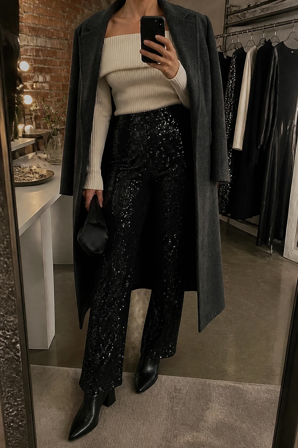 Image Prompt: Mirror selfie of a woman wearing an ivory fitted off-shoulder ribbed knit sweater with a wide folded neckline tucked into black high-waisted straight-leg sequin trousers, black leather pointed-toe ankle boots with block heels, a long charcoal grey tailored wool coat worn open, a small black satin clutch, and delicate silver stud earrings, holding a phone in front of a mirror, cropped at the NECK so the face does not show, with a relaxed casual selfie pose, in a fashion studio mirror corner, warm moody lighting, a clothing rack with garments behind and a jewelry tray on the vanity, realistic black sequins with subtle natural sparkle, soft ivory ribbed knit texture and understated evening styling, latest iPhone photo quality, no additional clothing or accessories.