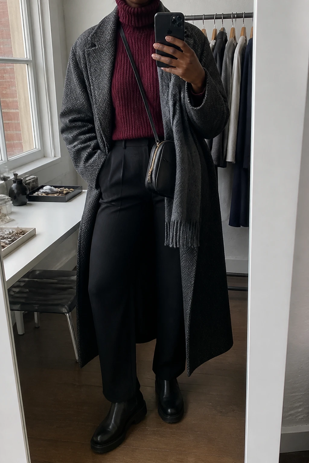 Image Prompt: Mirror selfie of a black woman wearing a long charcoal grey herringbone wool coat over a burgundy thick ribbed turtleneck sweater, black high-waisted straight-leg wool trousers, black waterproof leather chunky Chelsea boots with rubber soles, a structured black leather zippered crossbody bag, and a grey cashmere scarf, holding a phone in front of a mirror, cropped at the NECK so the face does not show, with a relaxed casual selfie pose, in a fashion studio mirror corner, soft natural window light, a clothing rack with garments behind and a jewelry tray on the vanity, realistic herringbone fabric, natural burgundy knit texture and subtle winter lighting, latest iPhone photo quality, no additional clothing or accessories.