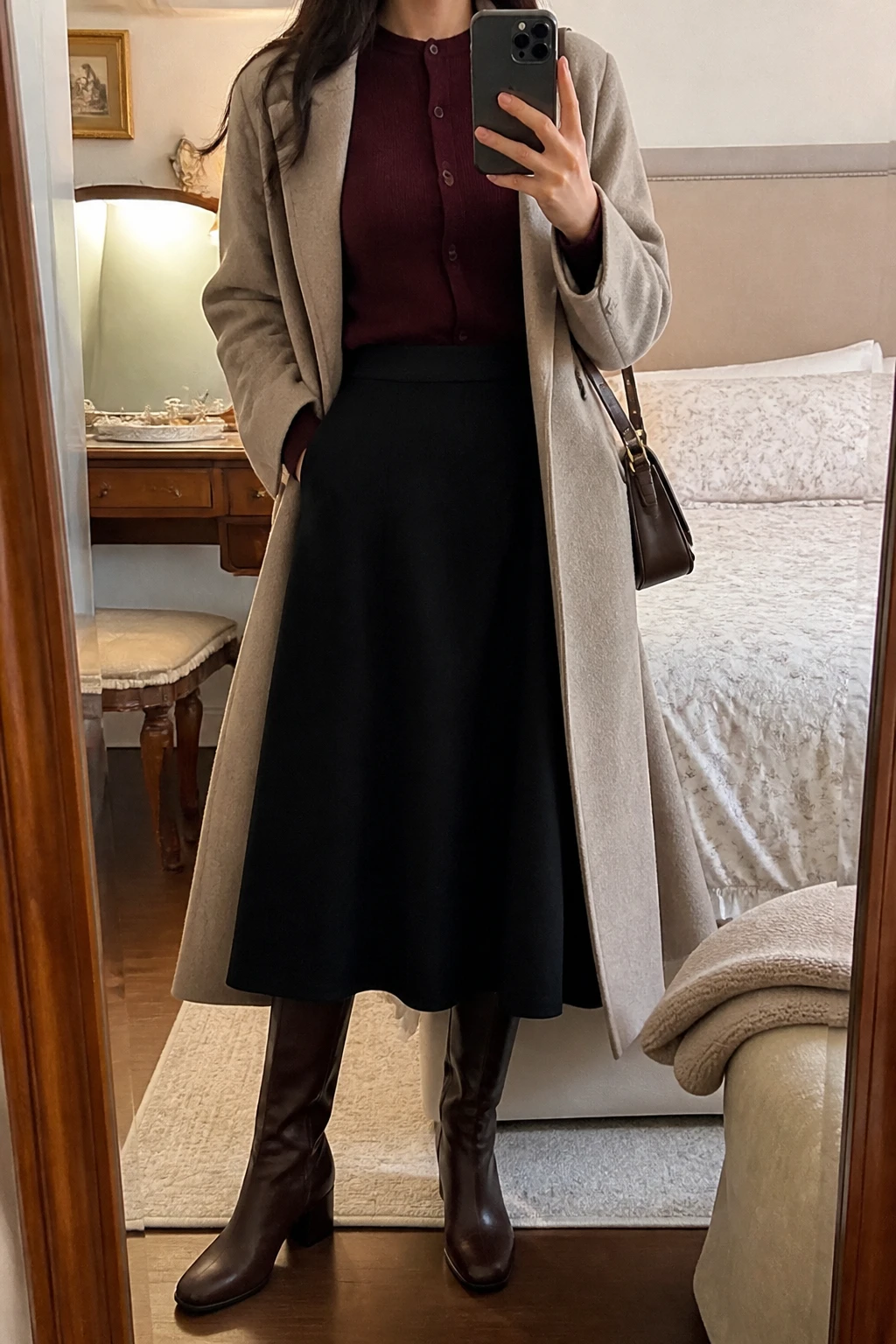 Image Prompt: Mirror selfie of a woman wearing a deep burgundy fine-knit wool button-front cardigan, fully buttoned and tucked into a black high-waisted A-line wool midi skirt, chocolate brown leather knee-high boots with low block heels, a long beige tailored wool coat worn open, and a structured dark brown leather shoulder bag, holding a phone in front of a mirror, cropped at the NECK so the face does not show, with a relaxed casual selfie pose, in a feminine-themed bedroom, warm soft indoor lighting, a folded blanket on a chair and a jewelry tray on the vanity, realistic burgundy wool knit texture, natural black A-line skirt folds and warm winter colors, latest iPhone photo quality, no additional clothing or accessories.