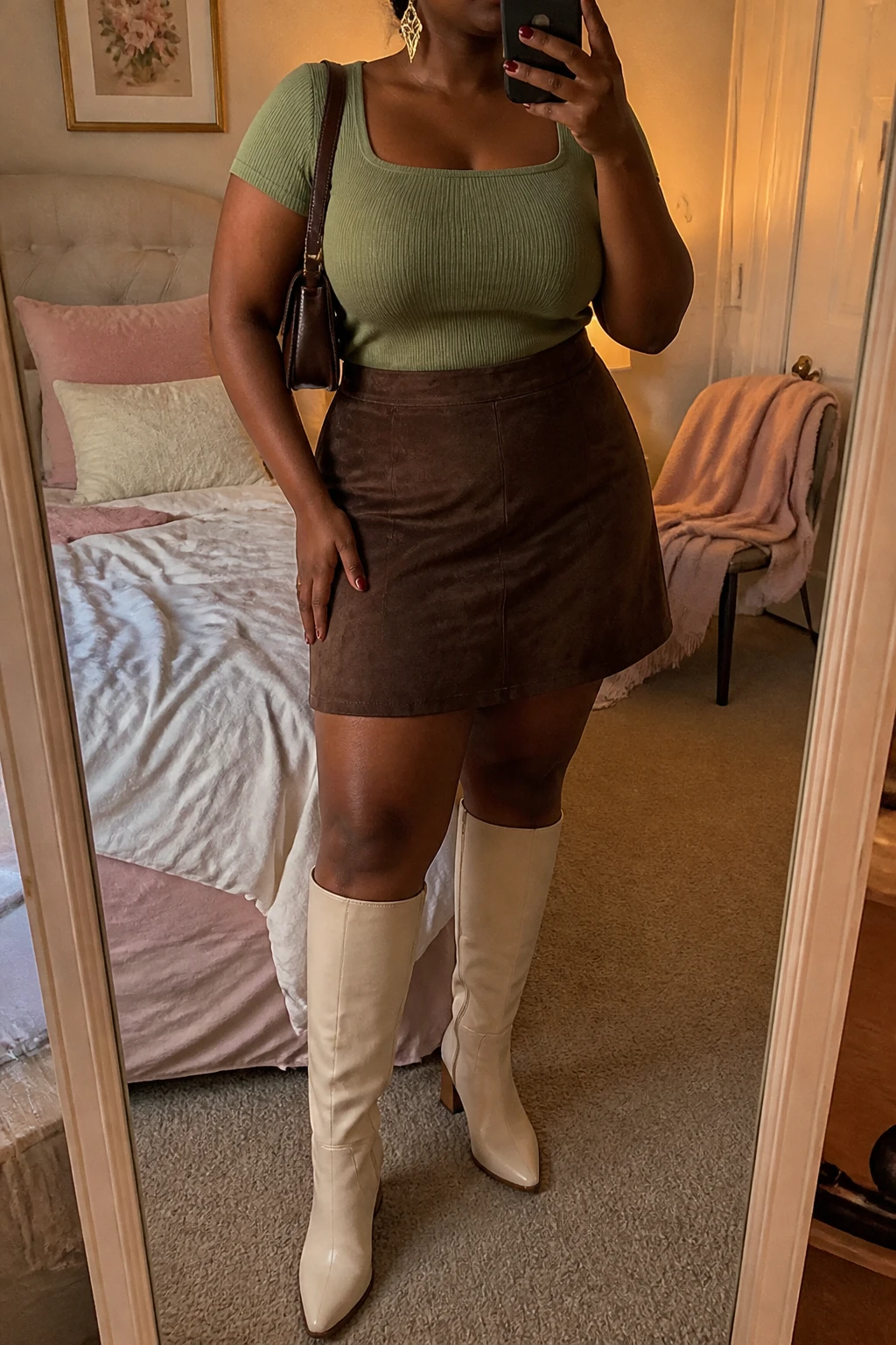Mirror selfie of a black plus size woman wearing a chocolate brown suede A-line mini skirt, a fitted sage green short-sleeve knit top with a square neckline, cream knee-high boots with low block heels, a small chocolate brown shoulder bag, and delicate gold drop earrings, clearly showing the exact clothing items, chocolate suede, sage knit, colors, footwear, and accessories listed, holding a phone in front of a mirror, cropped at the neck so her face is hidden, relaxed feminine selfie pose, in a feminine-themed bedroom with evening golden-hour lighting, soft pillows on an unmade bed and a folded blanket on a chair, realistic soft autumn styling, latest iPhone photo quality.