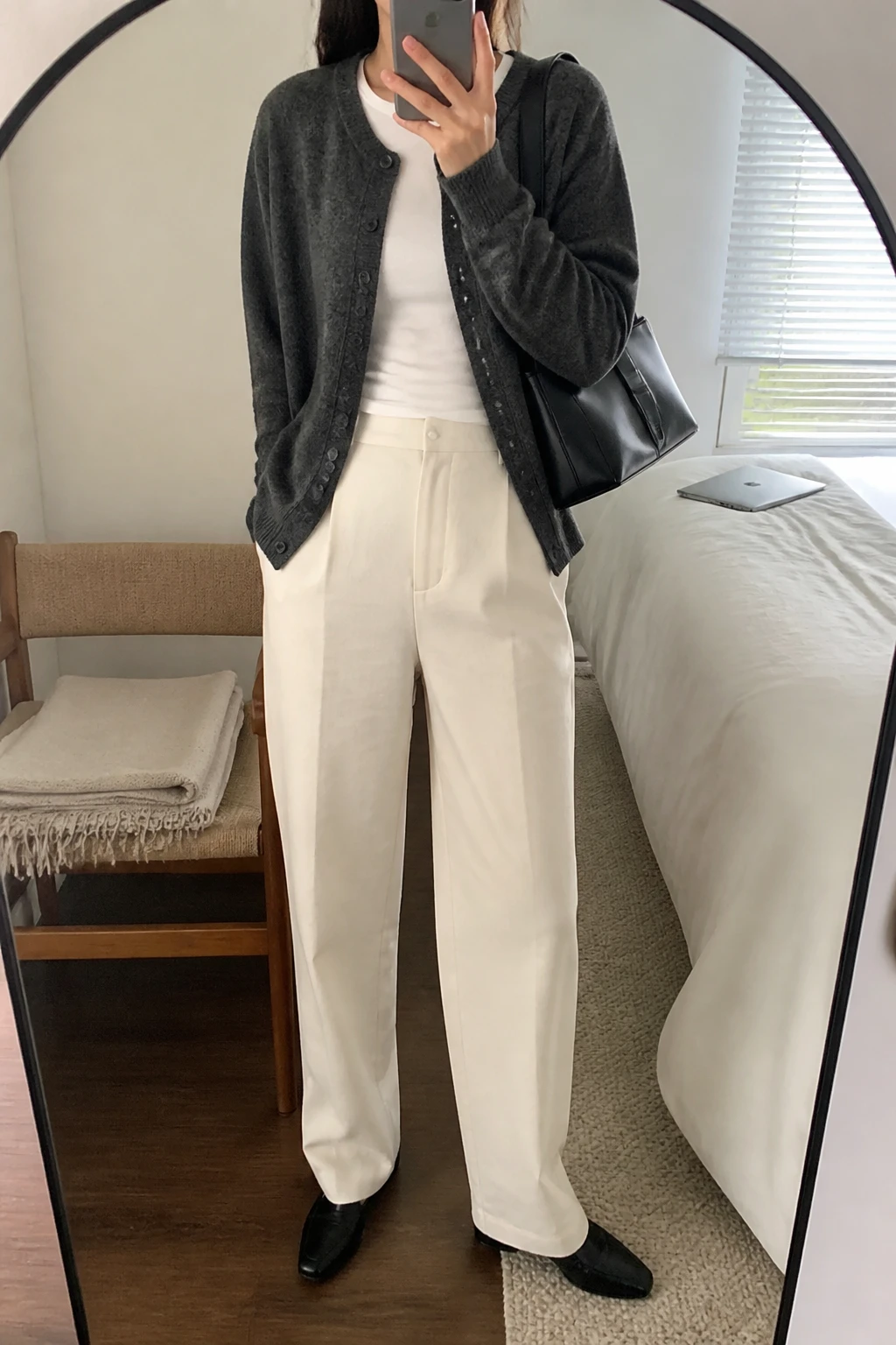Image Prompt: Mirror selfie of a woman wearing a charcoal grey fine-knit button-front cardigan over a white fitted cotton crewneck top, cream high-waisted straight-leg wool trousers, black leather pointed-toe loafers, and a medium structured black leather shoulder bag, holding a phone in front of a mirror, cropped at the NECK so the face does not show, with a relaxed casual selfie pose, in a cozy bedroom mirror setup, soft natural window light, a folded blanket on a chair and a laptop on the bed, realistic knit texture and natural trouser folds, latest iPhone photo quality, no additional clothing or accessories.