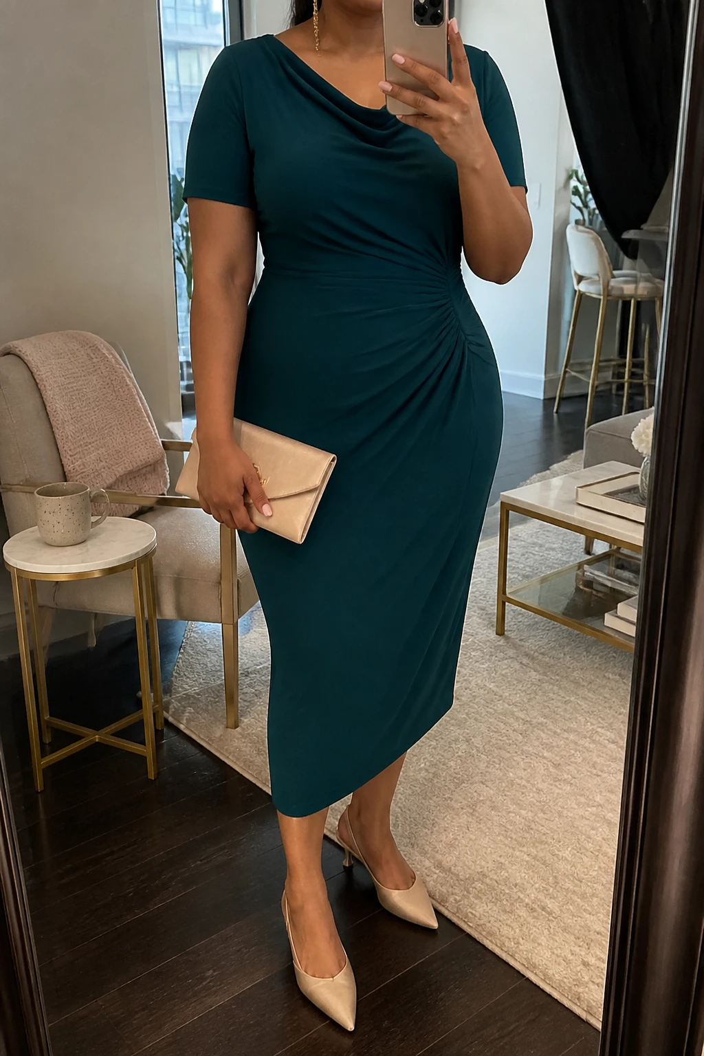Mirror selfie of a plus size woman wearing a teal green crepe midi dress with short sleeves, a softly draped neckline, and gentle gathering at one side of the waist, champagne pointed kitten heels, a champagne satin envelope clutch, and delicate gold chain earrings, clearly showing the exact dress, teal color, draping, footwear, and accessories listed, holding a phone in front of a mirror, cropped at the neck so the face does not show, relaxed elegant selfie stance, in a stylish city apartment with soft indoor lighting, a coffee mug on a side table and a folded blanket on a chair, realistic refined birthday lunch styling, latest iPhone photo quality.
