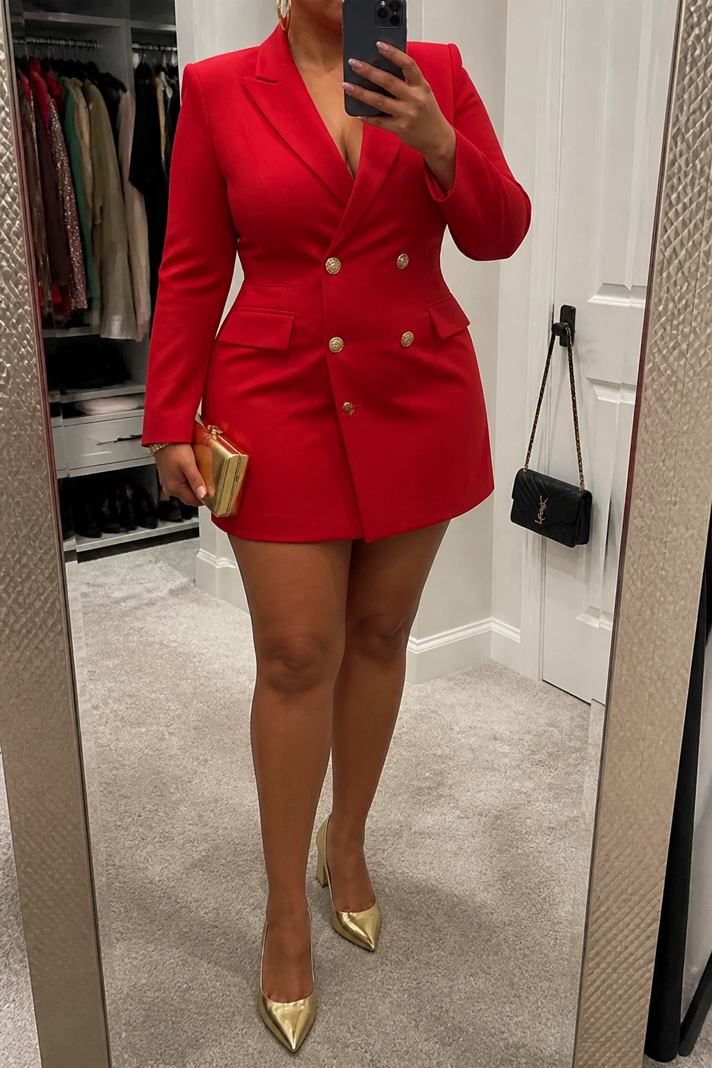Mirror selfie of a plus size woman wearing a structured red double-breasted blazer mini dress with long sleeves and a defined waist, metallic gold pointed pumps with low block heels, a small gold box clutch, and chunky gold hoop earrings, clearly showing the exact blazer dress, red color, gold footwear, and accessories listed, holding a phone in front of a mirror, cropped at the neck so her face does not appear, relaxed powerful selfie stance, in a walk-in closet mirror with soft indoor lighting, an open closet with clothes visible and a handbag hanging on a door handle, realistic birthday party styling, latest iPhone photo quality.