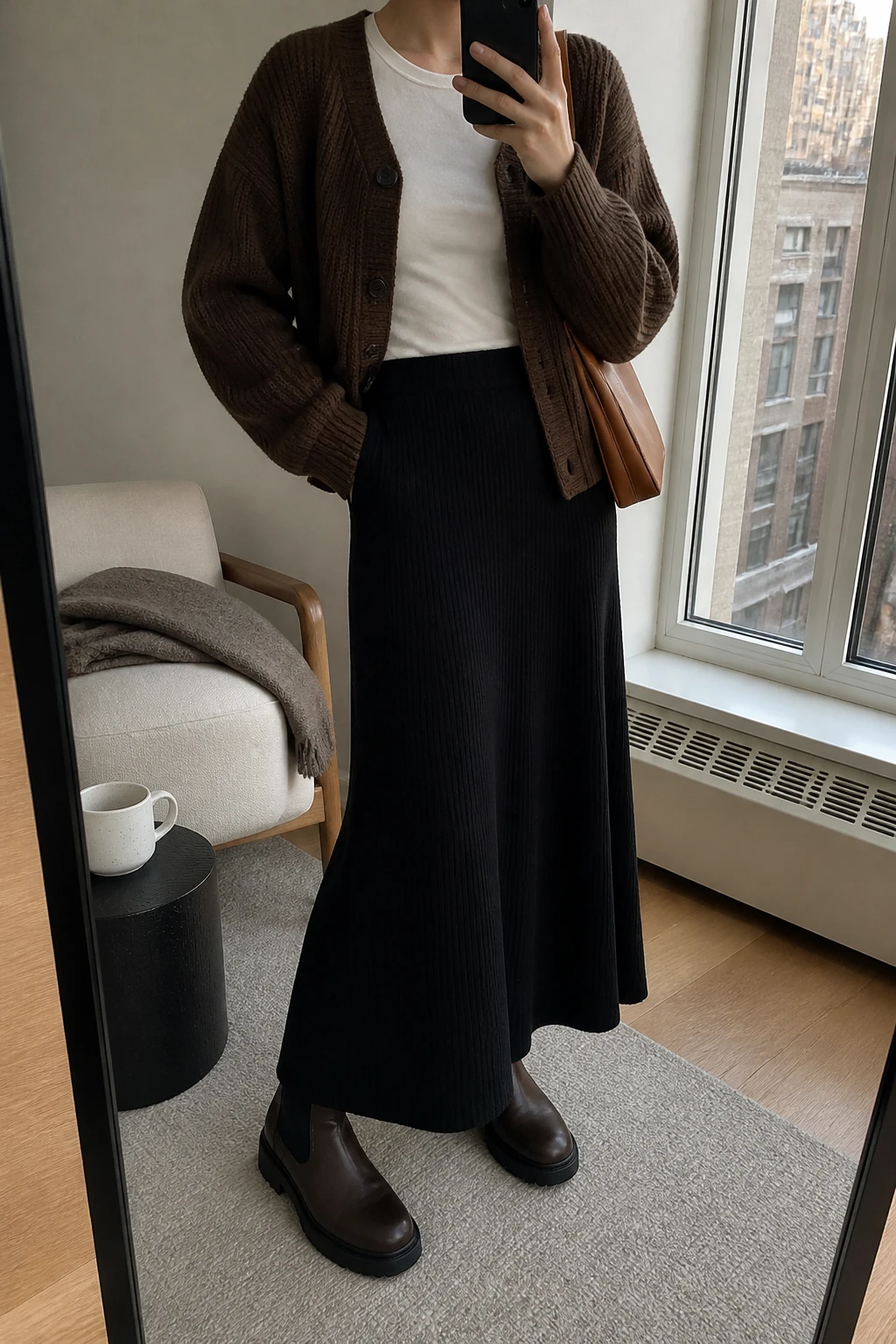 Image Prompt: Mirror selfie of a woman wearing a chocolate brown chunky-knit button-front cardigan over an ivory fitted long-sleeve cotton crewneck top, a black high-waisted ribbed knit A-line maxi skirt, dark brown leather chunky flat Chelsea boots, and a small caramel leather shoulder bag, holding a phone in front of a mirror, cropped at the NECK so the face does not show, with a relaxed casual selfie pose, in a stylish city apartment, soft natural window light, a coffee mug on a side table and a folded blanket on a chair, realistic chocolate cardigan stitches, natural black ribbed maxi skirt folds and soft warm-neutral colors, latest iPhone photo quality, no additional clothing or accessories.