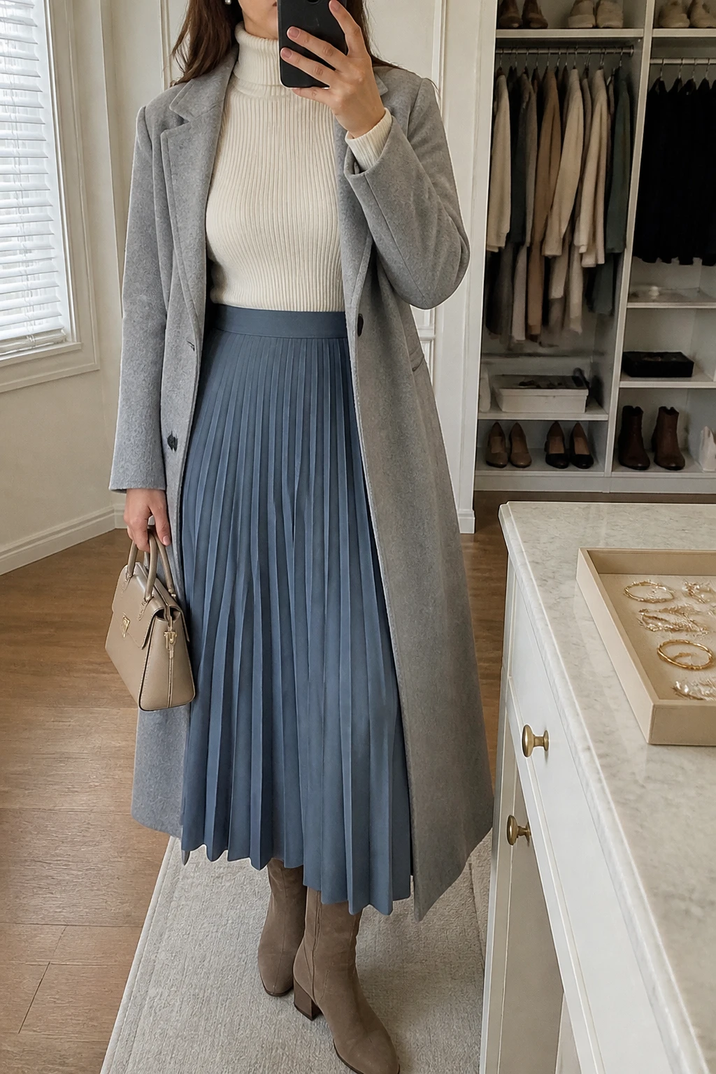 Image Prompt: Mirror selfie of a woman wearing an ivory fitted ribbed turtleneck sweater tucked into a dusty blue wool-blend pleated maxi skirt, taupe suede knee-high boots with low block heels, a long light grey tailored wool coat worn open, a small structured beige leather handbag, and delicate pearl stud earrings, holding a phone in front of a mirror, cropped at the NECK so the face does not show, with a relaxed casual selfie pose, in a walk-in closet mirror, soft natural window light, an open closet with clothes visible and a jewelry tray on the vanity, realistic dusty blue pleated wool texture, natural ivory ribbed knit and soft grey coat folds, latest iPhone photo quality, no additional clothing or accessories.