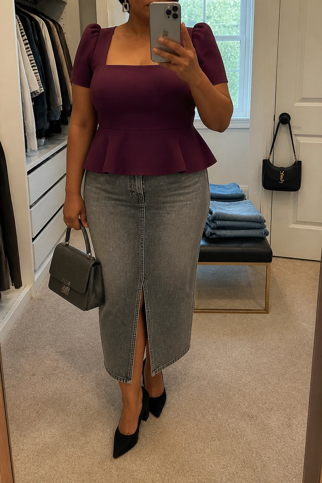 Mirror selfie of a plus size woman wearing a washed grey high-waisted denim midi skirt with a straight shape and front slit, a plum purple peplum top with a square neckline and short structured sleeves, black pointed block heels, a charcoal top-handle bag, and silver rectangular earrings, clearly showing the exact clothing items, grey denim, plum fabric, peplum shape, footwear, and accessories listed, holding a phone in front of a mirror, cropped at the neck so the face is hidden, relaxed polished selfie stance, in a walk-in closet mirror with natural window light, folded denim jeans on a bench and a handbag hanging on a door handle, realistic dinner outfit, latest iPhone photo quality.