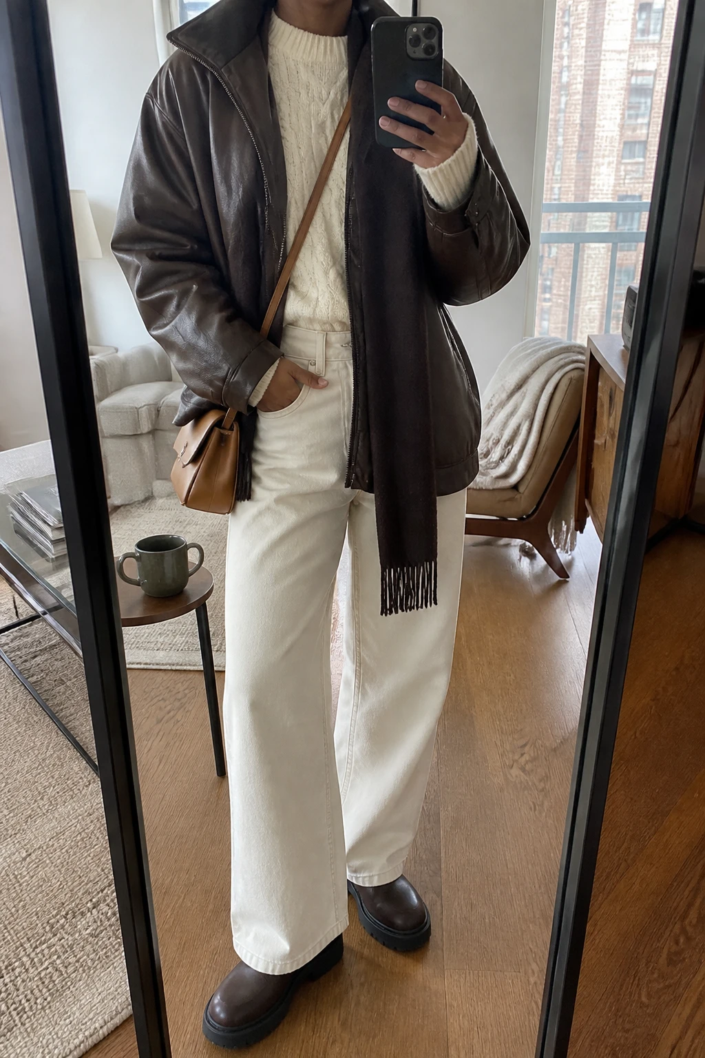 Image Prompt: Mirror selfie of a black woman wearing a chocolate brown oversized faux-leather jacket over a cream chunky cable-knit crewneck sweater, winter-white high-waisted wide-leg denim jeans, dark brown leather chunky flat ankle boots with rubber soles, a small tan leather crossbody bag, and a chocolate brown wool scarf, holding a phone in front of a mirror, cropped at the NECK so the face does not show, with a relaxed casual selfie pose, in a stylish city apartment, soft natural window light, a coffee mug on a side table and a folded blanket on a chair, realistic chocolate leather grain, natural white denim creases and soft cable-knit texture, latest iPhone photo quality, no additional clothing or accessories.