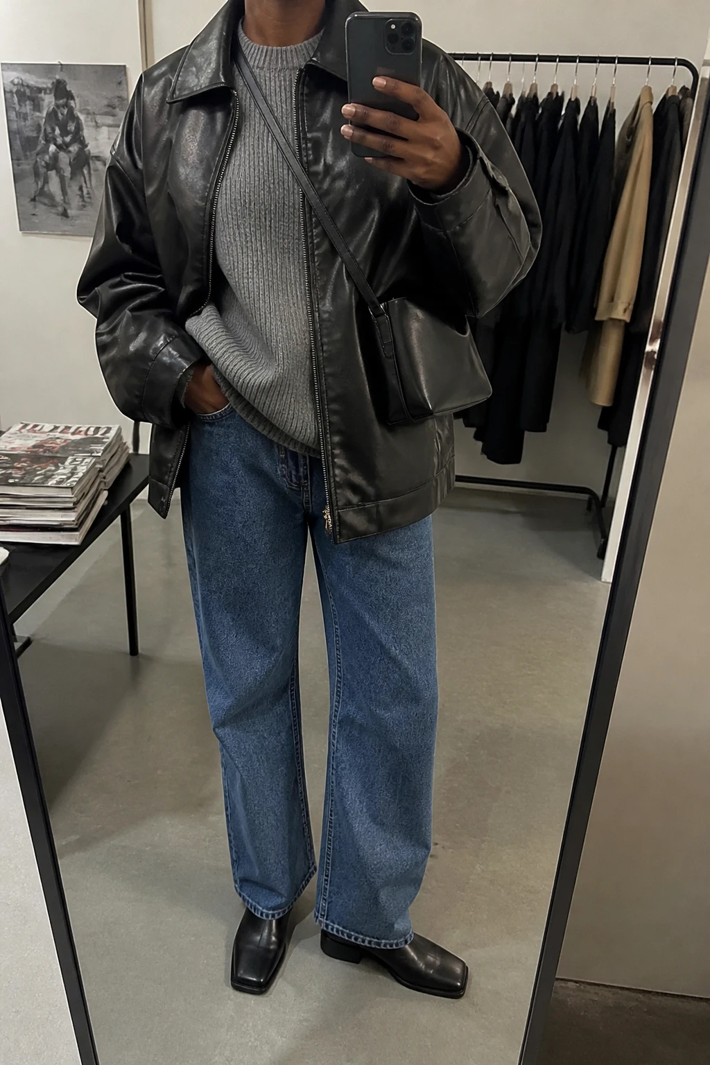 Image Prompt: Mirror selfie of a black woman wearing an oversized black faux-leather jacket over a heather grey thick ribbed crewneck sweater, medium blue relaxed straight-leg jeans, black leather square-toe flat ankle boots, and a small black leather crossbody bag, holding a phone in front of a mirror, cropped at the NECK so the face does not show, with a relaxed casual selfie pose, in a fashion studio mirror corner, soft indoor lighting, a clothing rack with garments behind and a stack of fashion magazines on a side table, realistic leather grain and knit texture, natural outfit folds, latest iPhone photo quality, no additional clothing or accessories.