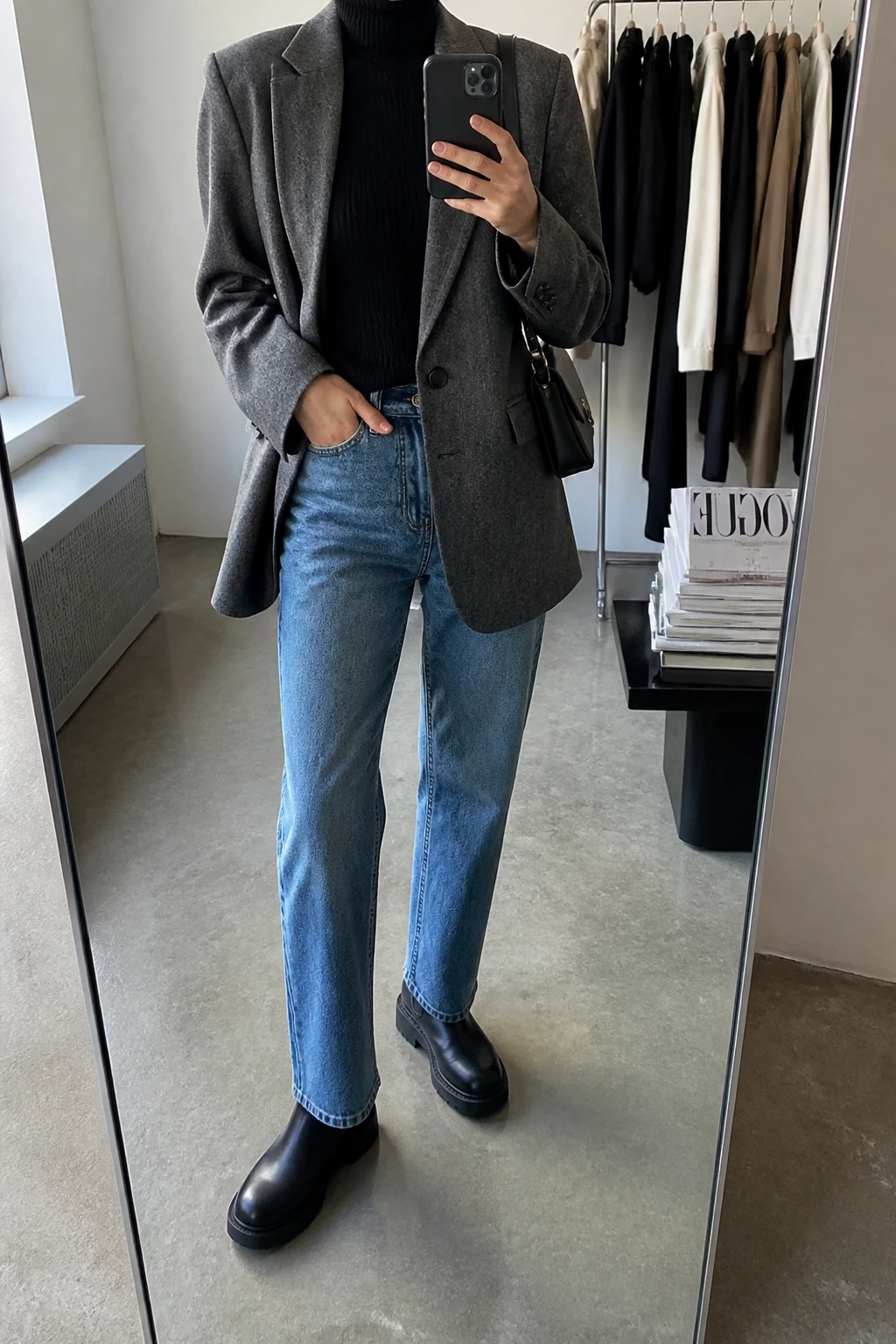 Image Prompt: Mirror selfie of a woman wearing an oversized heather grey wool-blend single-breasted blazer over a black fitted ribbed turtleneck sweater, medium-wash blue straight-leg jeans, black leather chunky flat Chelsea boots, and a small black structured shoulder bag, holding a phone in front of a mirror, cropped at the NECK so the face does not show, with a relaxed casual selfie pose, in a fashion studio mirror corner, soft natural window light, a clothing rack with garments behind and a stack of fashion magazines on a side table, realistic grey wool blazer texture and natural blue denim creases, latest iPhone photo quality, no additional clothing or accessories.