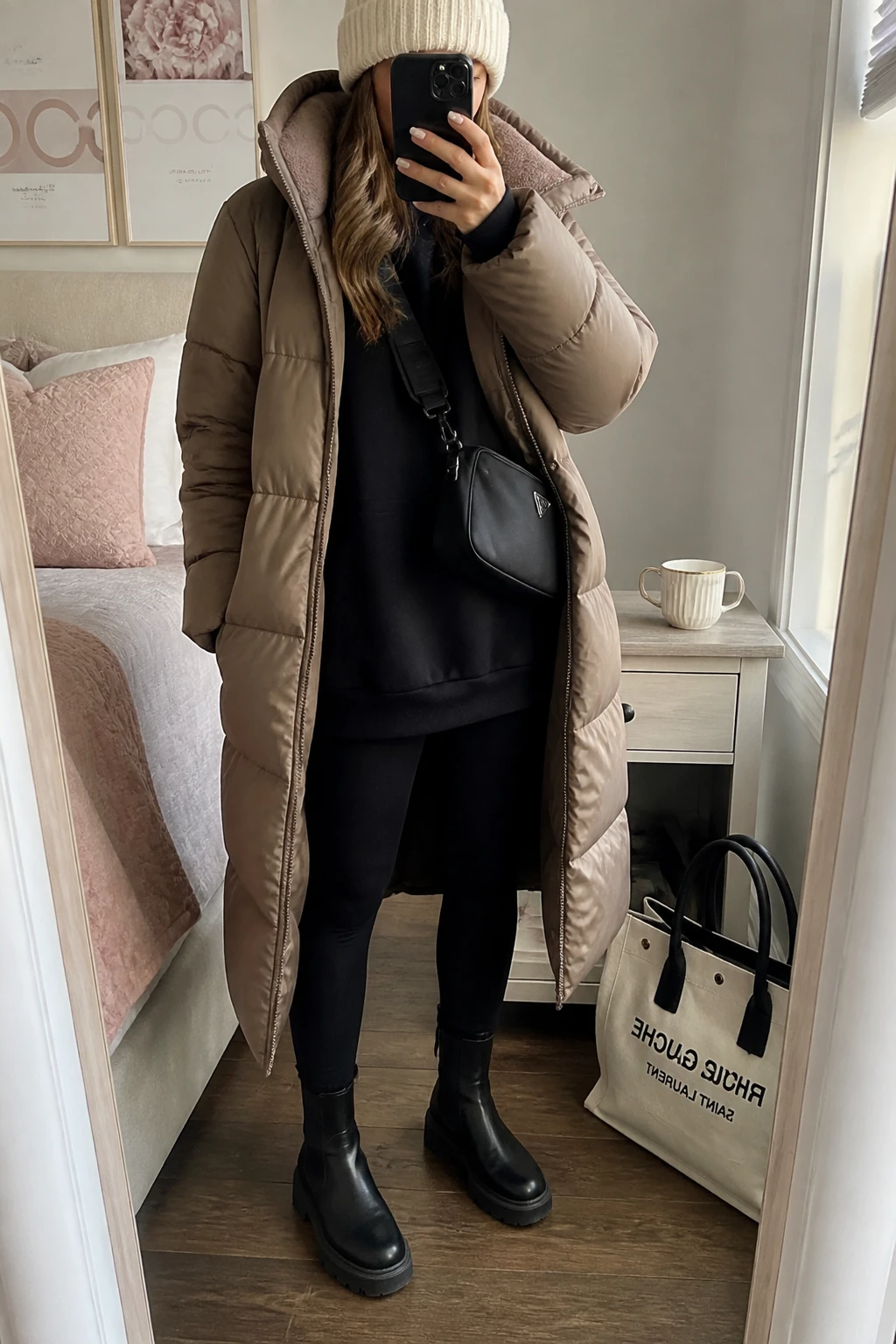 Image Prompt: Mirror selfie of a woman wearing a long taupe quilted down puffer coat over a black oversized fleece-lined sweatshirt, black thick thermal leggings, black waterproof leather ankle boots with flat lug soles, a black nylon crossbody bag, and a cream chunky ribbed knit beanie, holding a phone in front of a mirror, cropped at the NECK so the face does not show, with a relaxed casual selfie pose, in a feminine-themed bedroom, natural window light, a tote bag resting on the floor and a coffee mug on a bedside table, realistic winter coat volume, natural phone selfie framing, latest iPhone photo quality, no additional clothing or accessories.