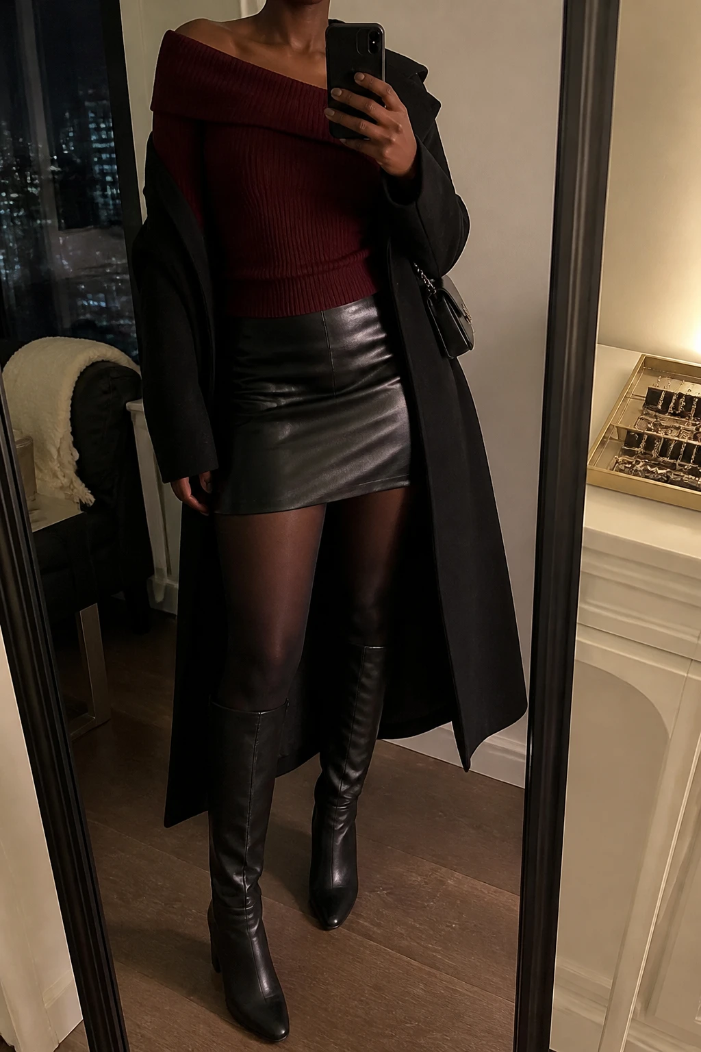 Image Prompt: Mirror selfie of a black woman wearing a deep burgundy fitted long-sleeve off-shoulder ribbed knit top with a wide folded neckline, a black high-waisted faux-leather mini skirt, black sheer-look fleece-lined thermal tights, black leather knee-high boots with sturdy block heels, a long black tailored wool coat worn open, and a small black chain-strap shoulder bag, holding a phone in front of a full-length mirror, cropped at the NECK so the face does not show, with a relaxed casual selfie pose, in a stylish city apartment, warm moody lighting, a jewelry tray on the vanity and a folded blanket on a chair, realistic burgundy knit texture, natural black leather grain, believable thermal tights and soft evening shadows, latest iPhone photo quality, no additional clothing or accessories.