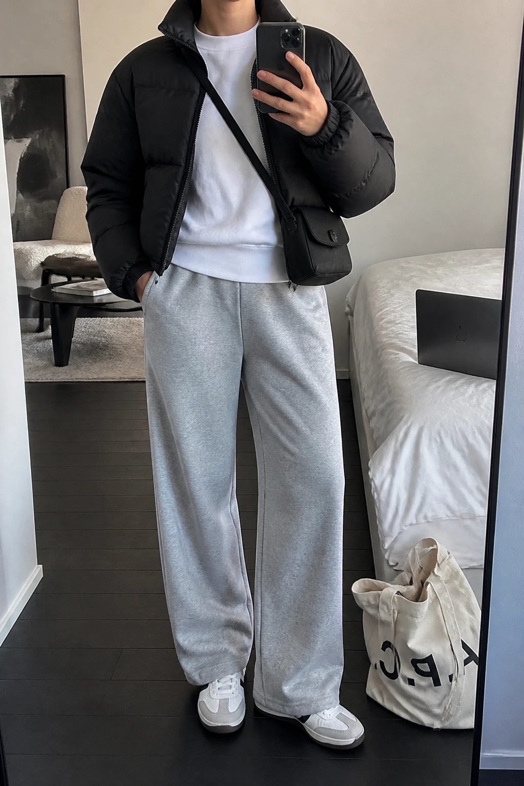 Image Prompt: Mirror selfie of a woman wearing a black cropped quilted down puffer jacket over a white heavyweight cotton crewneck sweatshirt, heather grey wide-leg fleece-lined sweatpants, white leather sneakers with grey detailing, and a small black nylon crossbody bag, holding a phone in front of a mirror, cropped at the NECK so the face does not show, with a relaxed casual selfie pose, in a stylish city apartment, soft indoor lighting, a tote bag resting on the floor and a laptop on the bed, natural puffer volume, realistic soft fleece fabric and casual proportions, latest iPhone photo quality, no additional clothing or accessories.