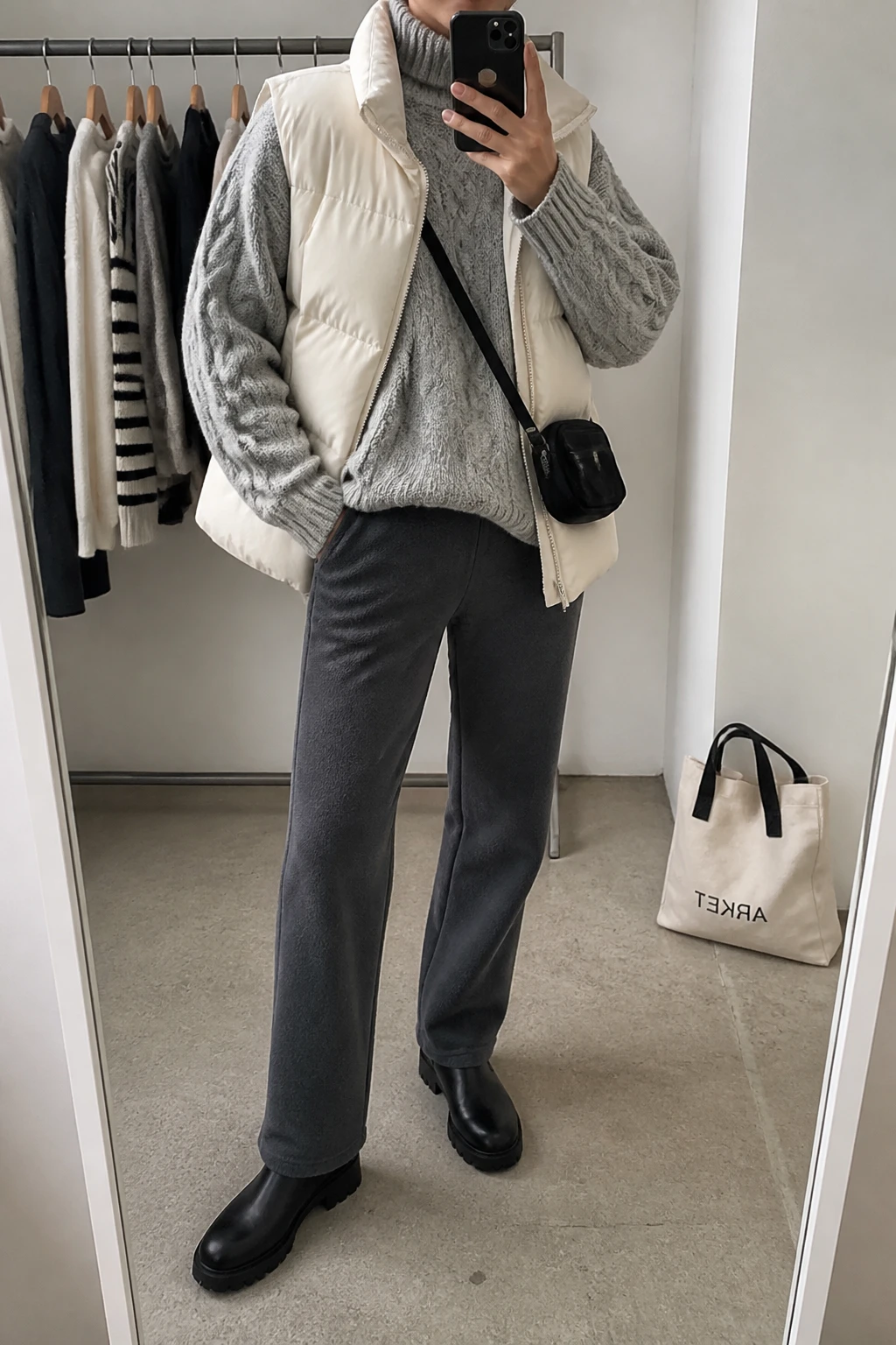 Image Prompt: Mirror selfie of a woman wearing a cream insulated quilted puffer vest over a heather grey chunky cable-knit turtleneck sweater, charcoal grey fleece-lined straight-leg trousers, black leather waterproof chunky ankle boots, and a small black nylon crossbody bag, holding a phone in front of a mirror, cropped at the NECK so the face does not show, with a relaxed casual selfie pose, in a fashion studio mirror corner, soft indoor lighting, a clothing rack with garments behind and a tote bag resting on the floor, realistic puffer fabric and chunky knit texture, latest iPhone photo quality, no additional clothing or accessories.