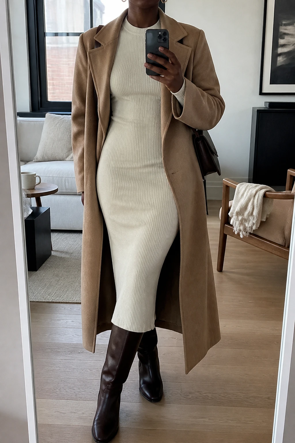 Image Prompt: Mirror selfie of a black woman wearing a cream long-sleeve ribbed knit midi dress with a softly fitted waist, chocolate brown leather knee-high boots with low block heels, a long camel wool wrap coat worn open, a small dark brown leather shoulder bag, and delicate gold hoop earrings, holding a phone in front of a full-length mirror, cropped at the NECK so the face does not show, with a relaxed casual selfie pose, in a stylish city apartment, soft natural window light, a coffee mug on a side table and a folded blanket on a chair, realistic ribbed knit texture and gentle wool coat folds, natural everyday appearance, latest iPhone photo quality, no additional clothing or accessories.