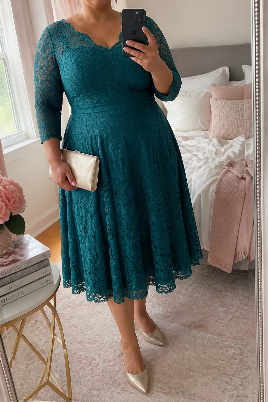 Mirror selfie of a plus size woman wearing a teal green lace midi dress with three-quarter sleeves, a scalloped neckline, a lined fitted bodice, and a softly flared skirt, champagne pointed kitten heels, a champagne satin clutch, and delicate gold stud earrings, clearly showing the exact clothing items, teal lace, lining, scalloped edges, footwear, and accessories listed, holding a phone in front of a mirror, cropped at the neck so her face does not show, relaxed graceful selfie pose, in a feminine-themed bedroom with natural window light, soft pillows on an unmade bed and a stack of fashion magazines on a side table, realistic graduation ceremony appearance, latest iPhone photo quality.