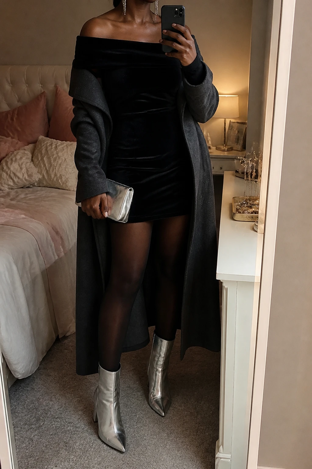Image Prompt: Mirror selfie of a black woman wearing a black long-sleeve off-shoulder velvet mini dress with a wide folded neckline and softly fitted waist, black sheer-look fleece-lined tights, silver metallic leather pointed-toe ankle boots with block heels, a long charcoal grey wool coat worn open, a small silver satin clutch, and delicate silver crystal drop earrings, holding a phone in front of a mirror, cropped at the NECK so the face does not show, with a relaxed casual selfie pose, in a feminine-themed bedroom, warm moody lighting, a jewelry tray on the vanity and soft pillows on an unmade bed, realistic black velvet texture, natural silver leather reflections, soft thermal tights finish, latest iPhone photo quality, no additional clothing or accessories.