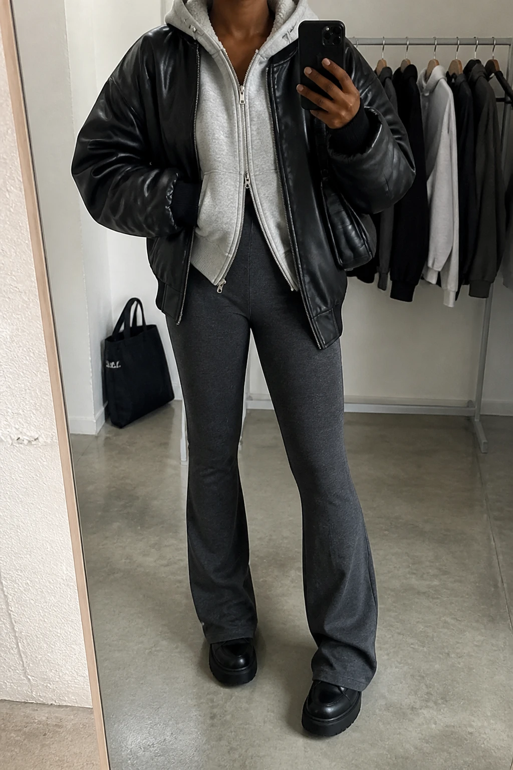 Image Prompt: Mirror selfie of a black woman wearing an oversized black faux-leather bomber jacket over a heather grey thick fleece-lined zip-up hoodie, charcoal grey high-waisted fleece-lined flared leggings, black chunky platform leather sneakers with flat rubber soles, and a small black nylon shoulder bag, holding a phone in front of a mirror, cropped at the NECK so the face does not show, with a relaxed casual selfie pose, in a fashion studio mirror corner, soft indoor lighting, a clothing rack with garments behind and a tote bag resting on the floor, realistic black leather grain, natural hoodie folds and soft charcoal leggings fabric, latest iPhone photo quality, no additional clothing or accessories.