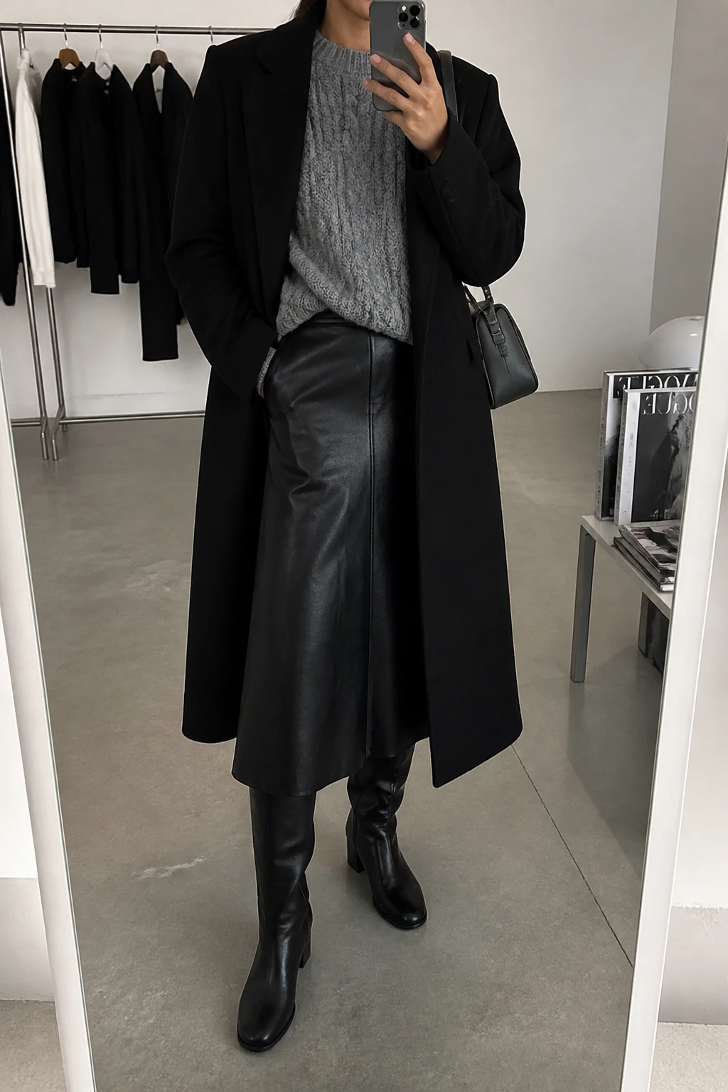 Image Prompt: Mirror selfie of a woman wearing a heather grey chunky cable-knit crewneck sweater with a small front tuck into a black high-waisted faux-leather A-line midi skirt, black leather knee-high boots with low block heels, a long black tailored wool coat worn open, and a small charcoal grey leather shoulder bag, holding a phone in front of a mirror, cropped at the NECK so the face does not show, with a relaxed casual selfie pose, in a fashion studio mirror corner, soft indoor lighting, a clothing rack with garments behind and a stack of fashion magazines on a side table, realistic grey cable-knit texture, natural black faux-leather grain and soft evening shadows, latest iPhone photo quality, no additional clothing or accessories.
