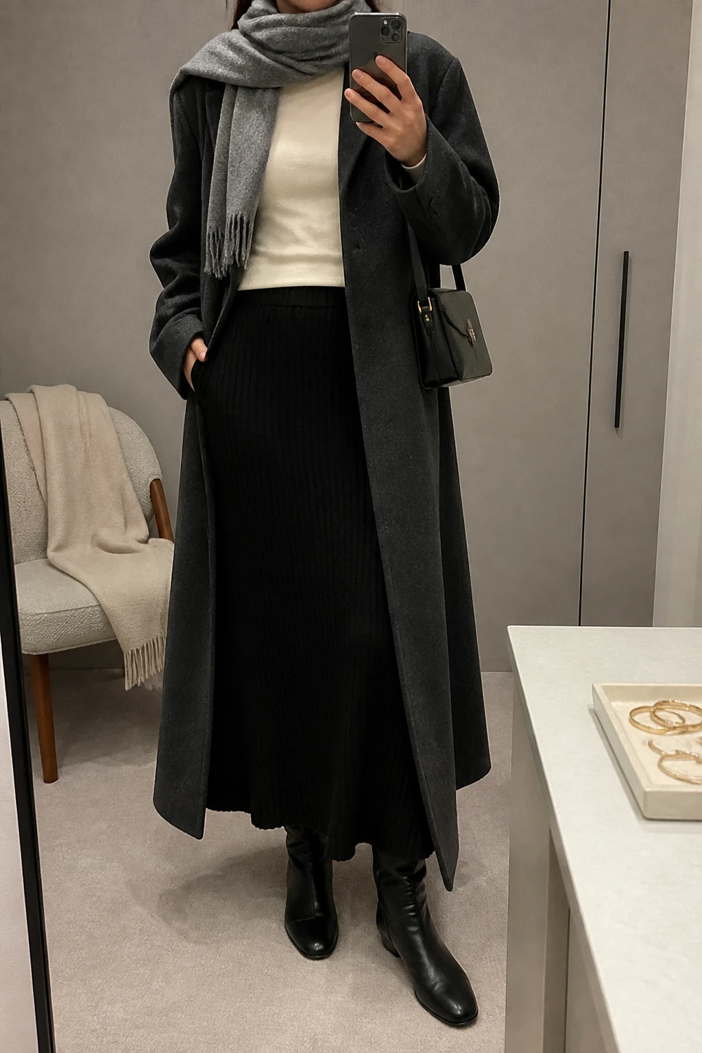 Image Prompt: Mirror selfie of a woman wearing a long charcoal grey oversized single-breasted wool coat over a cream fitted cashmere mock-neck sweater, a black high-waisted A-line ribbed knit maxi skirt, black leather knee-high flat boots, a small structured black leather shoulder bag, and a soft grey cashmere scarf wrapped neatly around the neck, holding a phone in front of a mirror, cropped at the NECK so the face does not show, with a relaxed casual selfie pose, in a minimal dressing room, soft indoor lighting, a folded blanket on a chair and a jewelry tray on the vanity, realistic charcoal wool coat texture, natural black ribbed skirt folds, soft cream cashmere fabric, latest iPhone photo quality, no additional clothing or accessories.