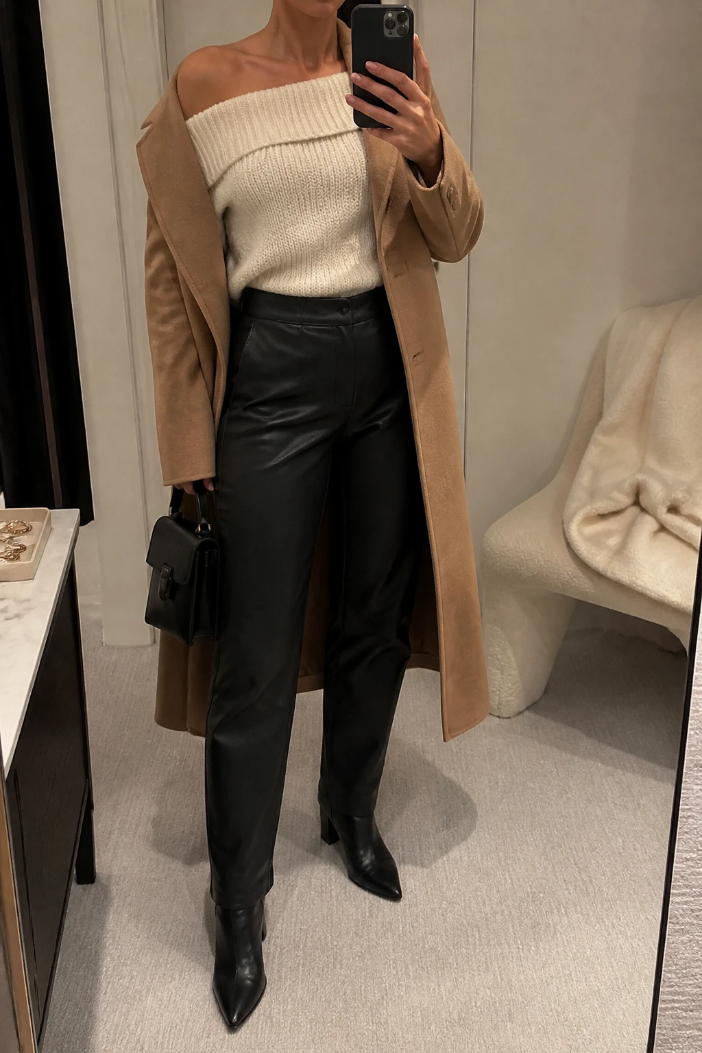 Image Prompt: Mirror selfie of a woman wearing a cream fitted off-shoulder chunky-knit sweater with a folded neckline, black high-waisted straight-leg faux-leather trousers, black leather pointed-toe ankle boots with block heels, a long camel tailored wool coat worn open, and a small structured black leather handbag, holding a phone in front of a mirror, cropped at the NECK so the face does not show, with a relaxed casual selfie pose, in a minimal dressing room, warm soft indoor lighting, a folded blanket on a chair and a jewelry tray on the vanity, realistic cream chunky-knit texture, natural black leather trouser folds, elegant winter evening styling, latest iPhone photo quality, no additional clothing or accessories.