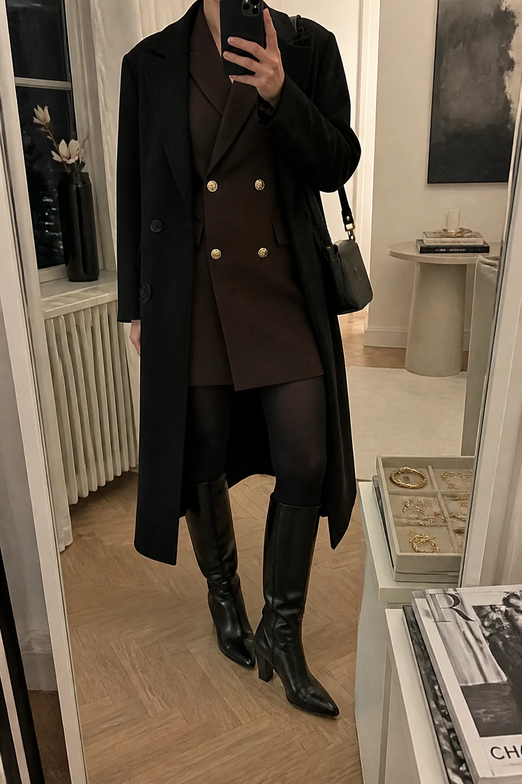 Image Prompt: Mirror selfie of a woman wearing a chocolate brown long-sleeve double-breasted wool-blend blazer mini dress with a defined waist and subtle gold buttons, black opaque fleece-lined tights, black leather knee-high boots with block heels, a long black wool coat worn open, and a small black leather shoulder bag, holding a phone in front of a mirror, cropped at the NECK so the face does not show, with a relaxed casual selfie pose, in a stylish city apartment, warm soft indoor lighting, a stack of fashion magazines on a side table and a jewelry tray on the vanity, realistic chocolate wool tailoring, natural black leather boots and refined evening styling, latest iPhone photo quality, no additional clothing or accessories.
