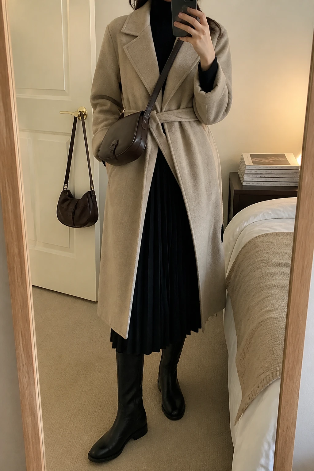Image Prompt: Mirror selfie of a woman wearing a long beige belted wool wrap coat loosely tied at the waist over a black fitted cashmere turtleneck sweater, a black wool-blend pleated midi skirt, black opaque thermal tights, black leather knee-high flat boots with rubber soles, and a small dark brown leather crossbody bag, holding a phone in front of a mirror, cropped at the NECK so the face does not show, with a relaxed casual selfie pose, in a cozy bedroom mirror setup, warm soft indoor lighting, a stack of fashion magazines on a side table and a handbag hanging on a door handle, realistic beige wool fabric, natural black skirt pleats and comfortable winter layering, latest iPhone photo quality, no additional clothing or accessories.